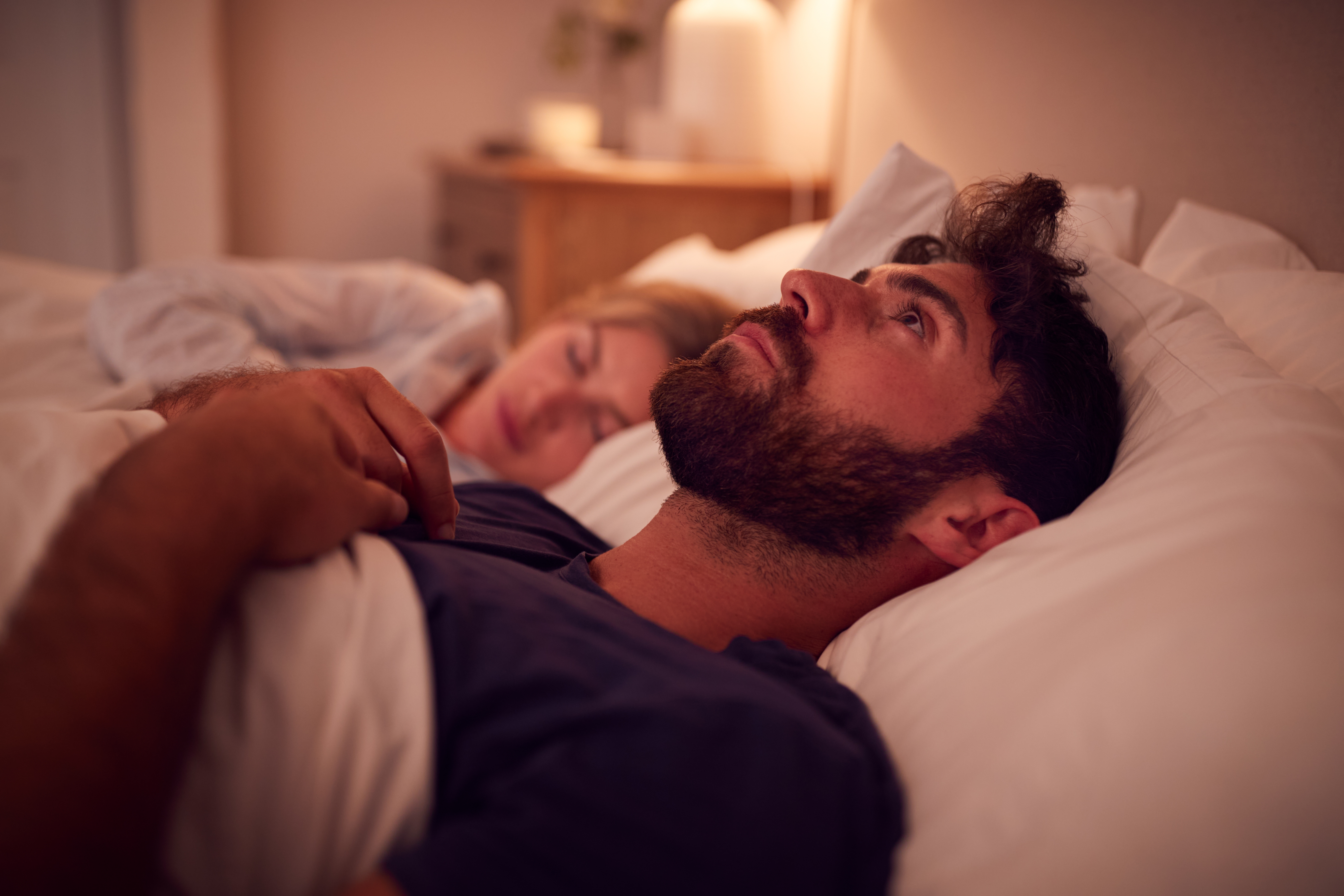 Man lying awake in bed, gazing upward, beside a sleeping woman. Cozy bedroom setting