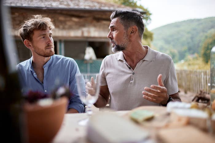 Two men sit at an outdoor table engaged in conversation, with one gesturing expressively, surrounded by blurred food and plants