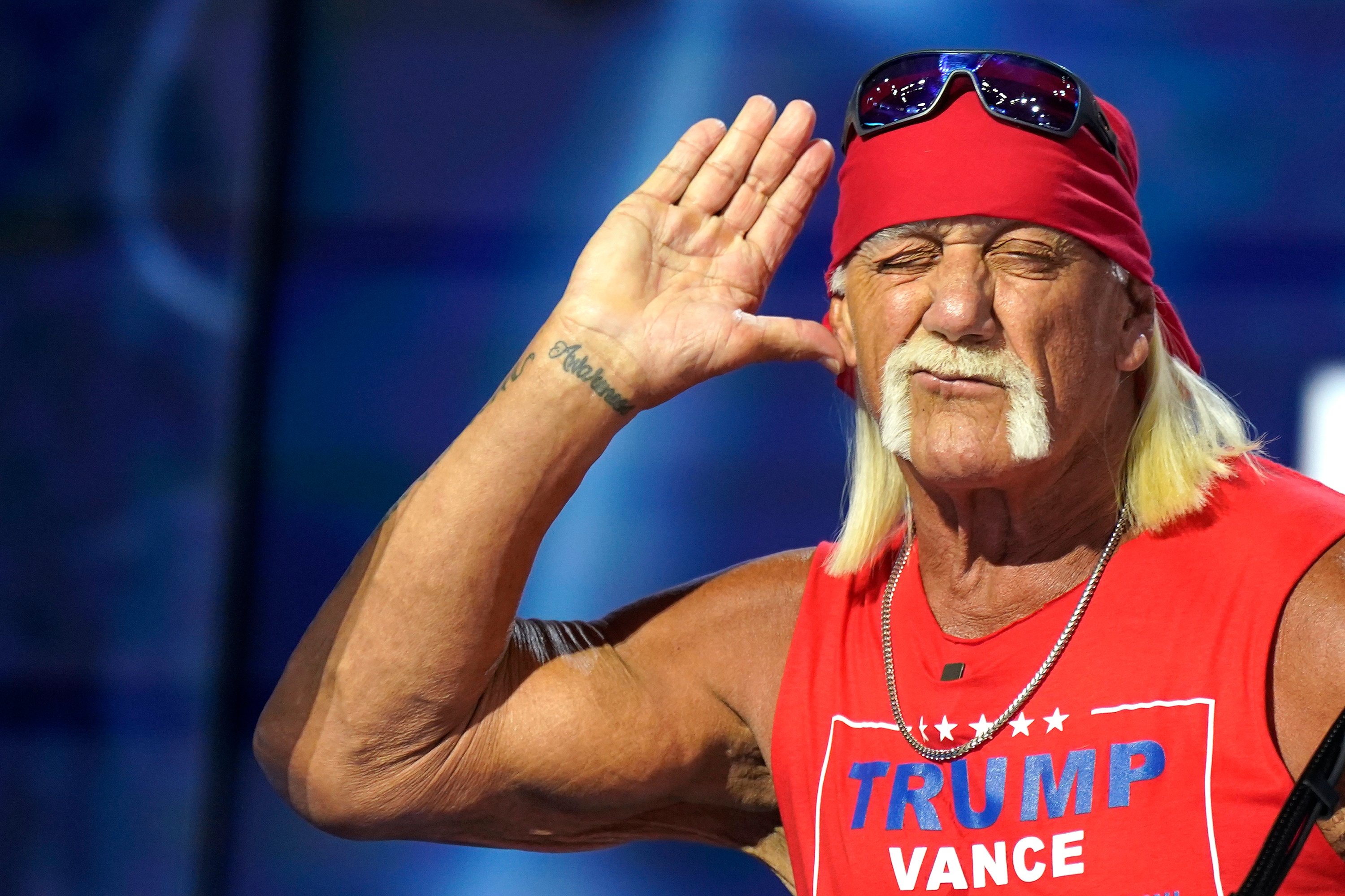 Hulk Hogan with long hair and mustache wearing a sleeveless top and bandana, holding hand to ear