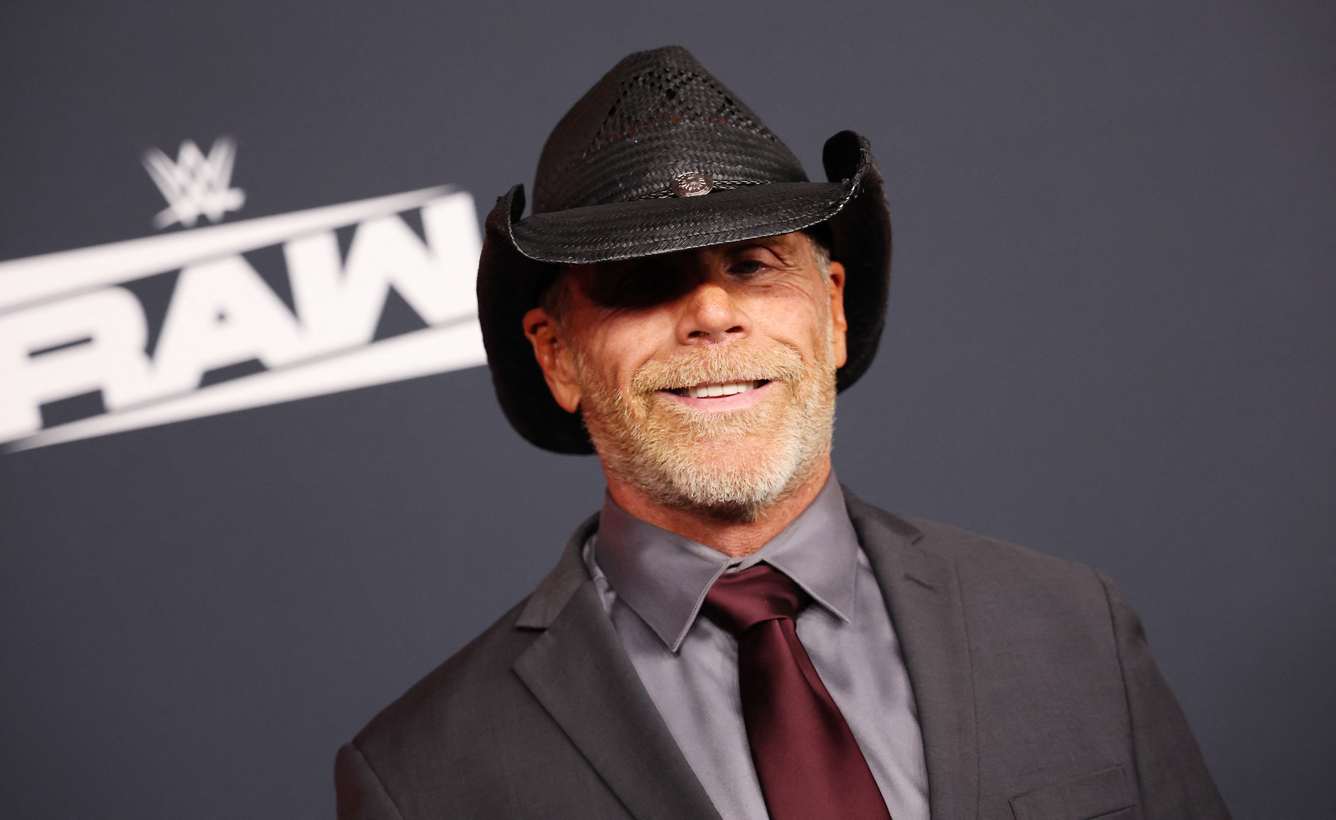 Shawn Michaels in a cowboy hat and suit with tie, smiling at a WWE event backdrop