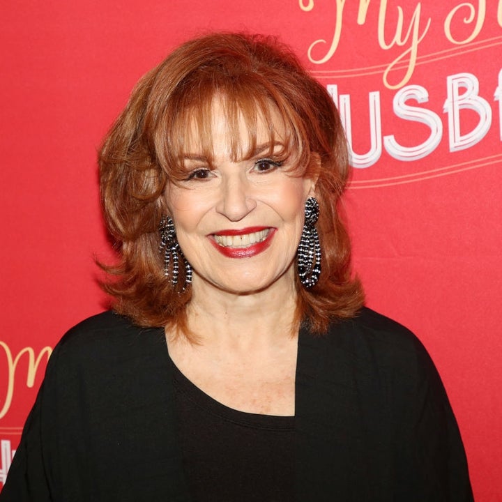 Person smiling at an event with a red backdrop, wearing a black outfit and statement earrings