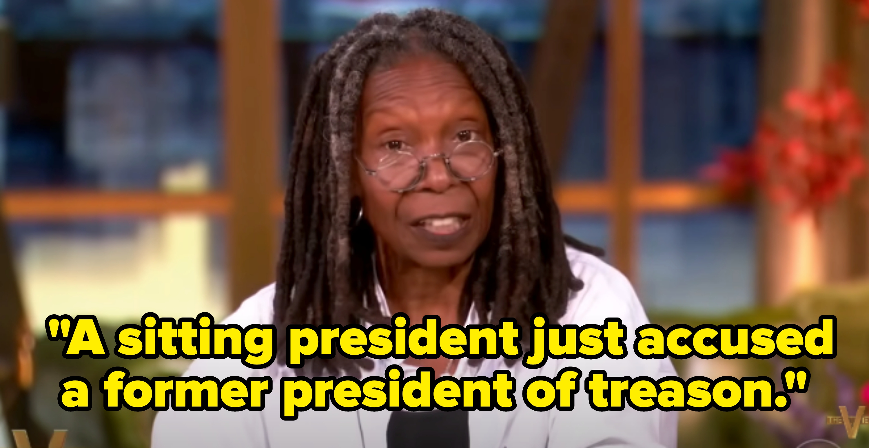 Person with long braided hair and round glasses, wearing a white shirt, speaks on a talk show set