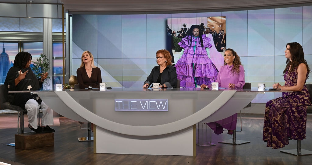 Five hosts sit behind a semi-circular table on a TV talk show set; a display screen shows a large photo behind them