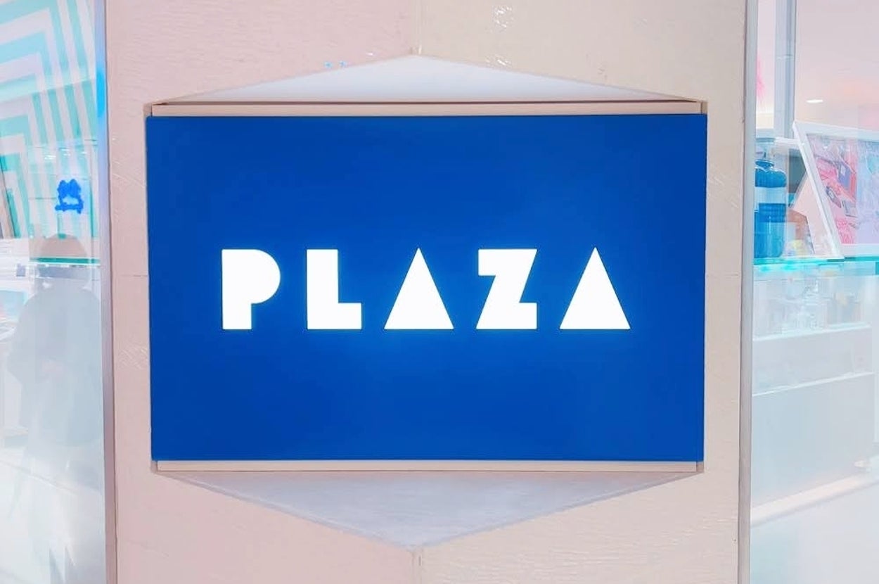 「PLAZA」と書かれた看板が壁にはめ込まれている。周囲には商品が陳列された棚が見える。