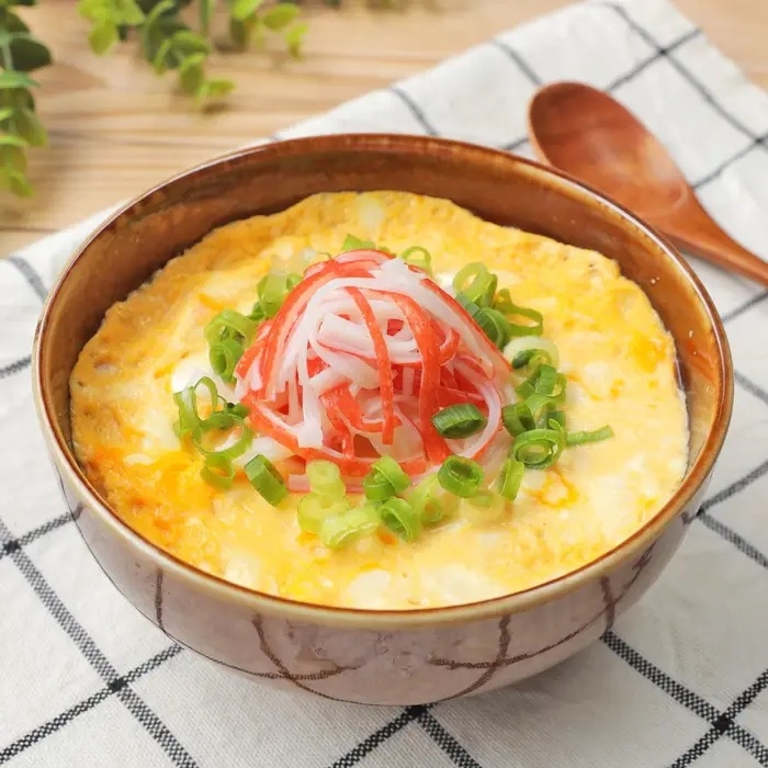 かにかま、ネギを乗せた卵丼がテーブルに置かれている。背景には緑の植物と木製のスプーン。