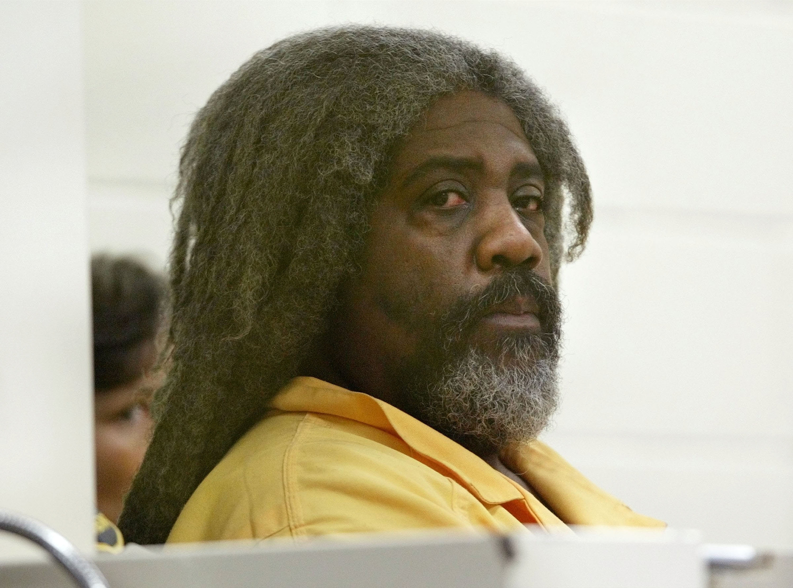 I don’t know who this is, but a man with long hair and a beard is sitting in what appears to be a courtroom, wearing a beige suit