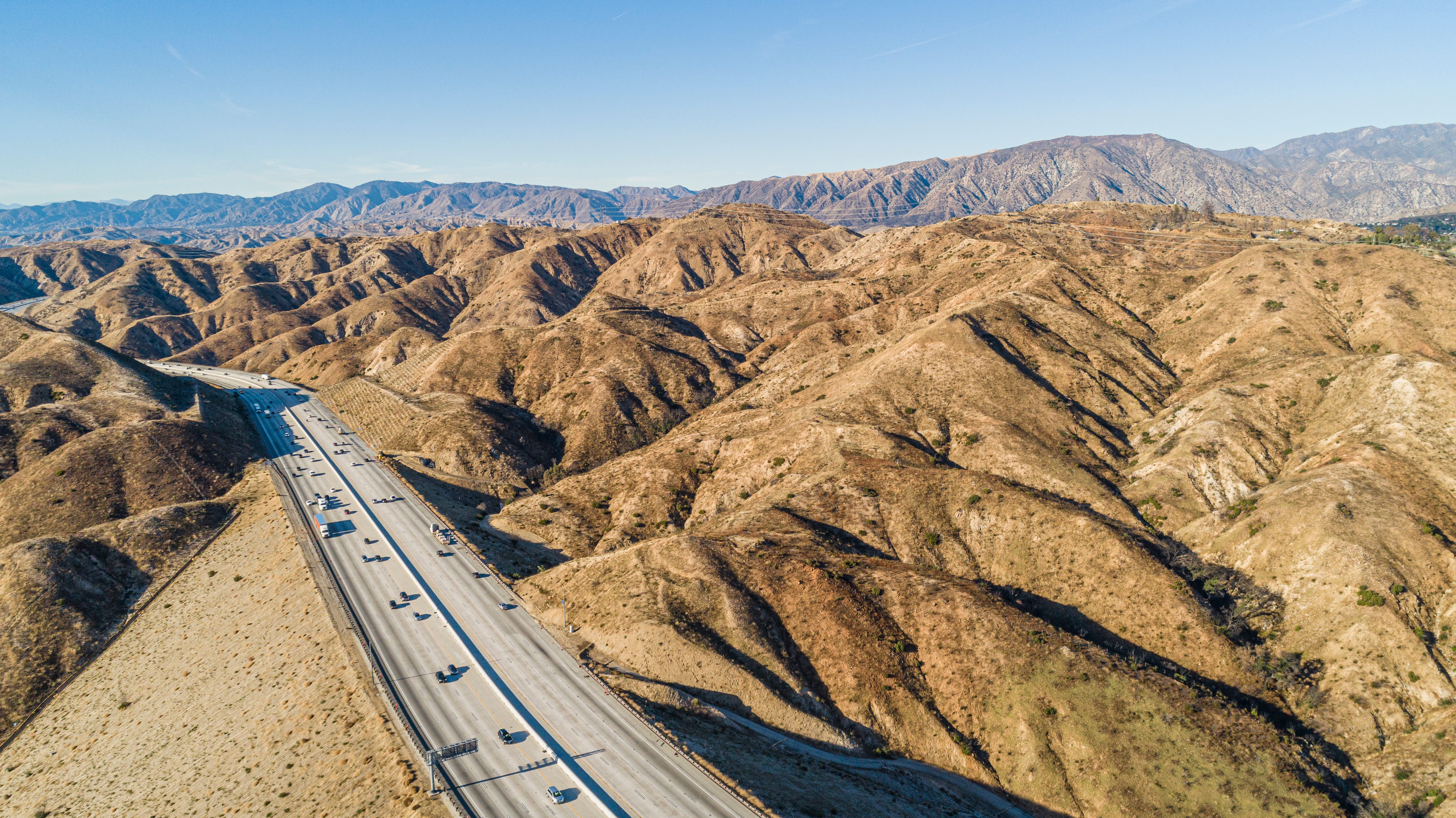 Aerial view of a highway cutting through arid, hilly terrain with mountains in the background, illustrating a scenic, expansive landscape