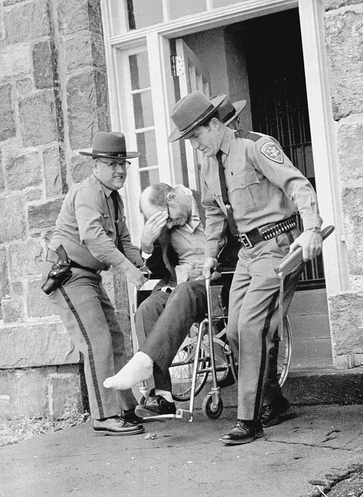 Two police officers assist a man in a wheelchair, helping him exit a building
