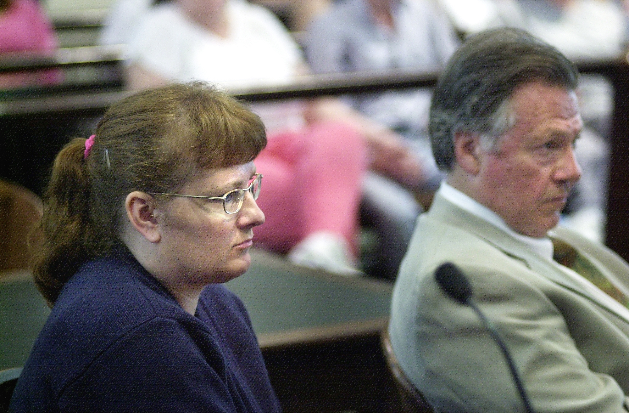 Two people seated attentively in a courtroom setting, audience visible in the background