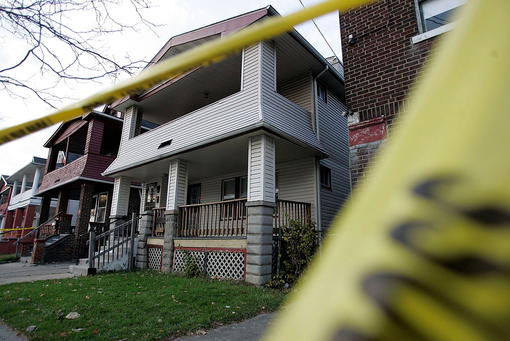 House behind yellow caution tape, suggesting a restricted area or investigation scene. Residential neighborhood with similar houses visible