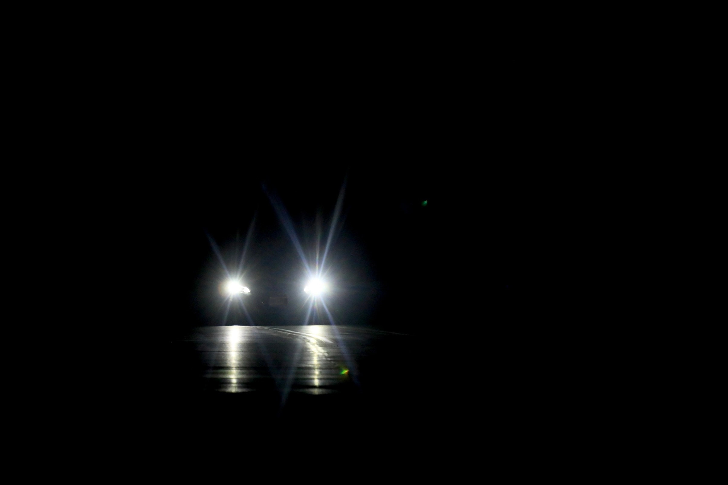 Two car headlights shine brightly in a darkened setting, illuminating the area directly ahead on what appears to be a nighttime road