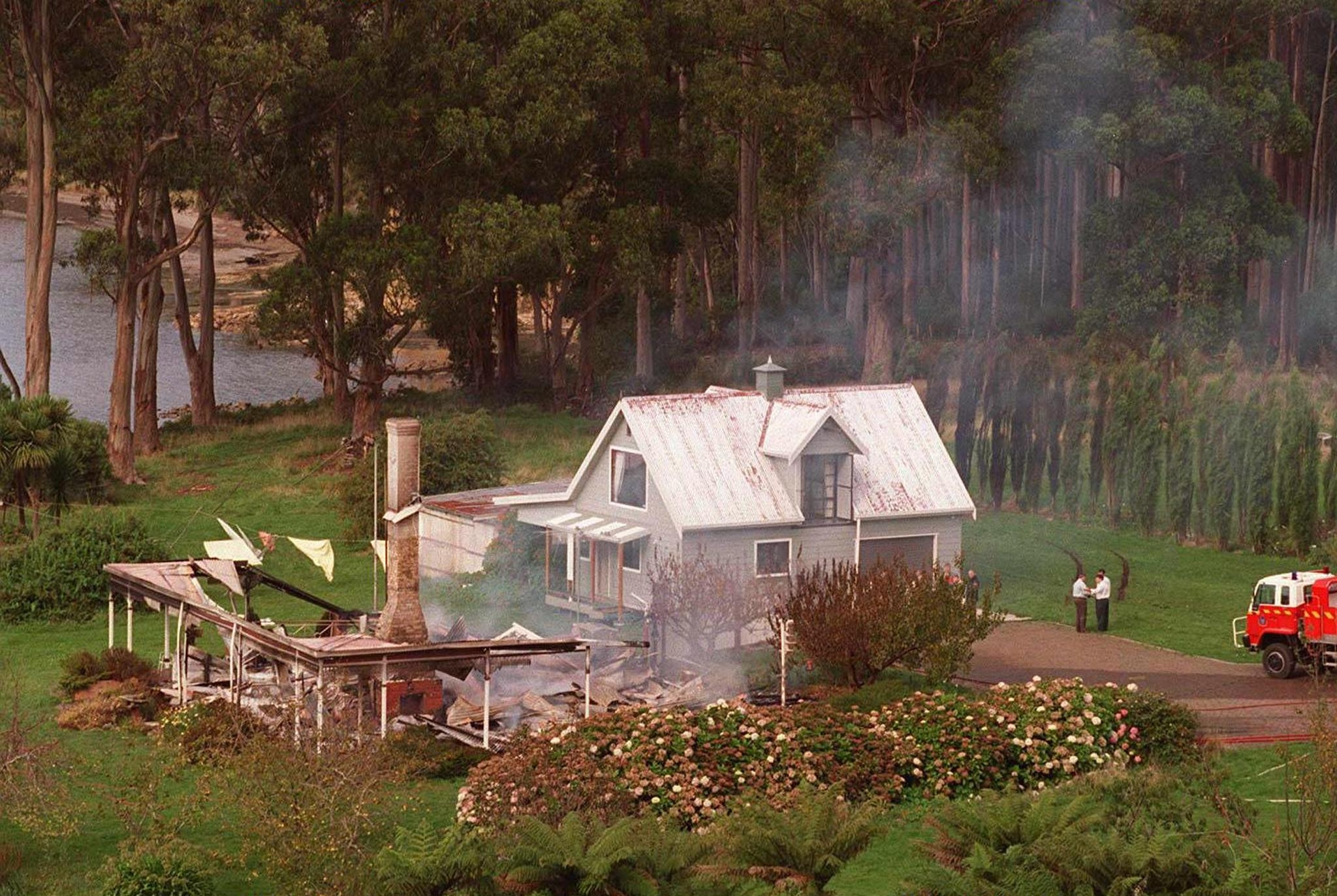 A quaint house with a damaged roof beside a smoking structure. Two people stand nearby, and a fire truck is parked on a driveway. Forest in the background