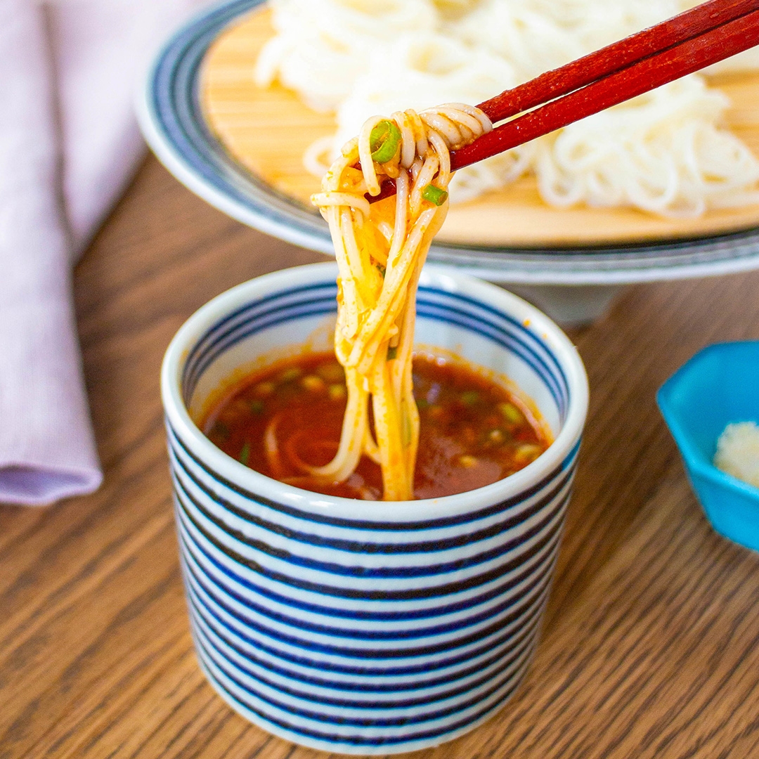 麺が箸で持ち上げられ、たれに浸かっている。奥に麺の皿が見える。