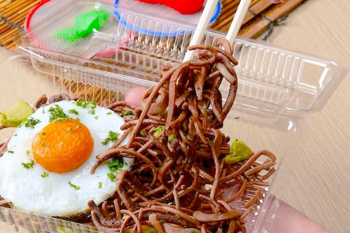 箸で持ち上げたそばと目玉焼き、わさびの付いた弁当。背景にカラフルなスプーンと絵の具。