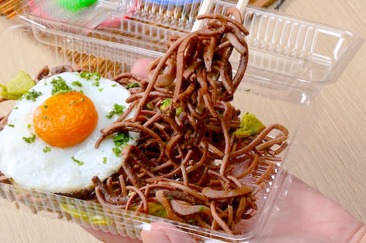 箸で持ち上げたそばと目玉焼き、わさびの付いた弁当。背景にカラフルなスプーンと絵の具。