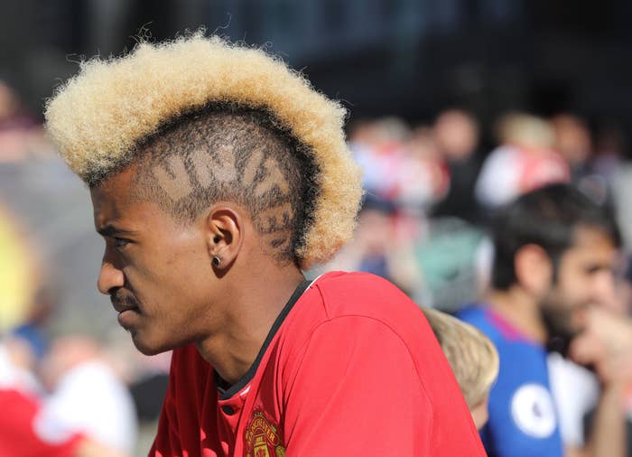 Person with a blonde mohawk, shaved sides featuring a design, wearing a sports jersey with the Manchester United logo