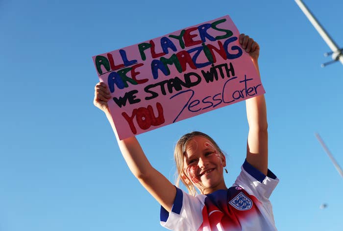 Child holding a sign that says, "All players are amazing. We stand with you, Jess Carter," wearing an England soccer jersey