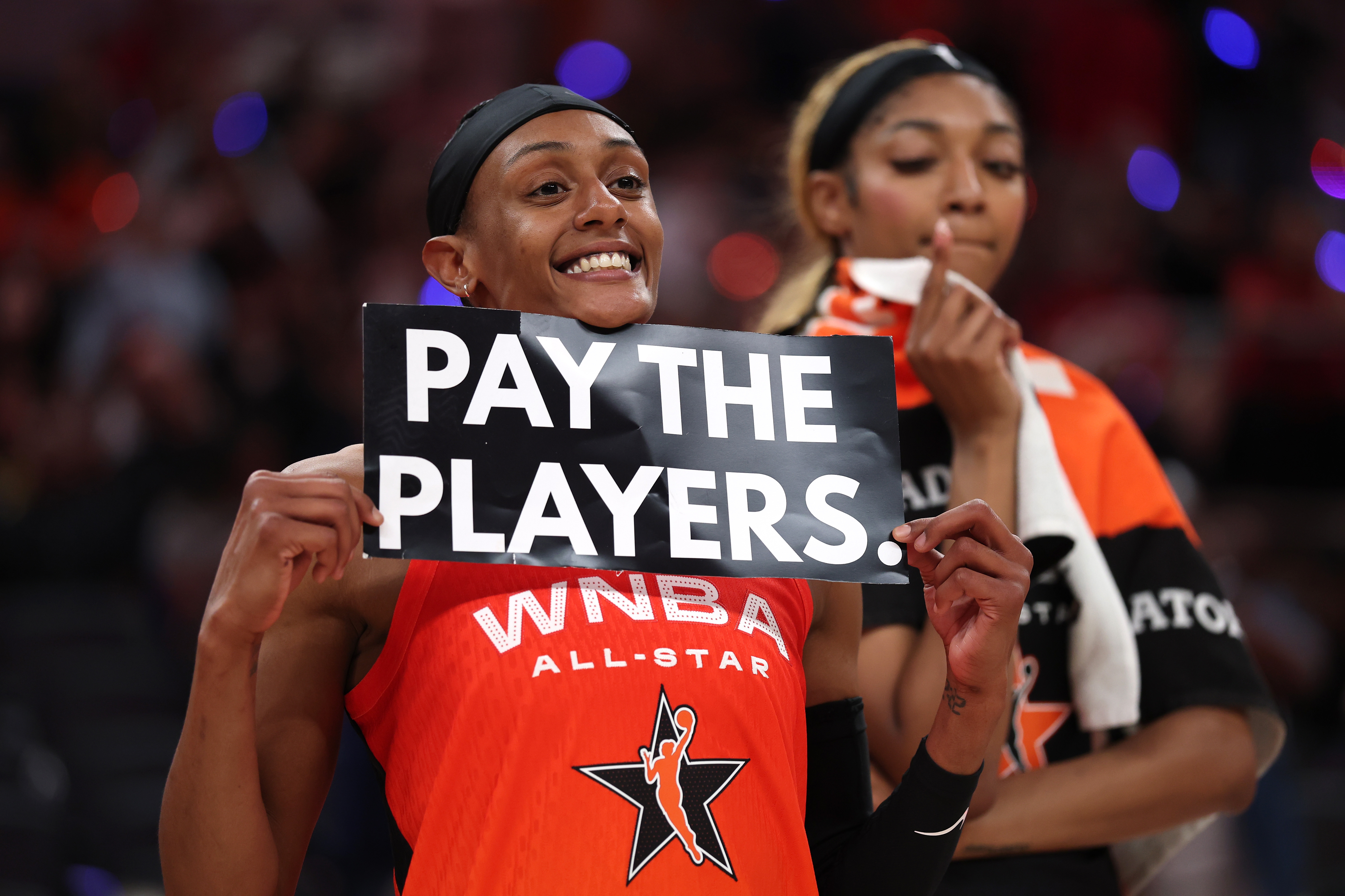 WNBA player holds "Pay the Players" sign at an All-Star event, advocating for equitable compensation in sports. Another player stands in the background
