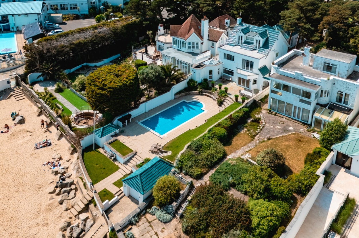 Aerial view of a spacious beachside estate with multiple white buildings, a central swimming pool, and lush gardens near the sandy shore