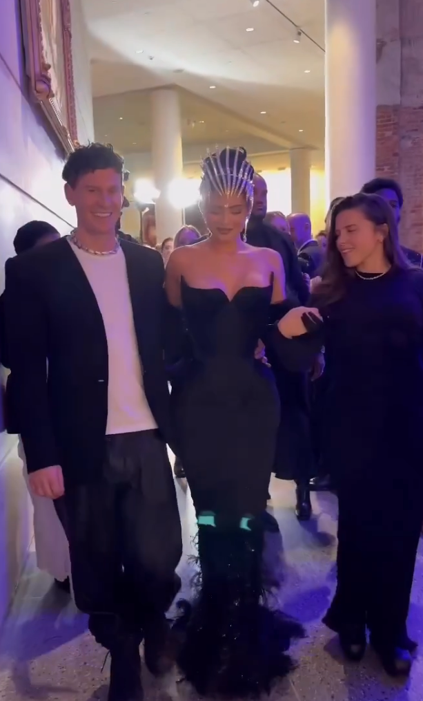 Kylie in an elegant off-the-shoulder gown with ornate headpiece, walking supported by two people at an event