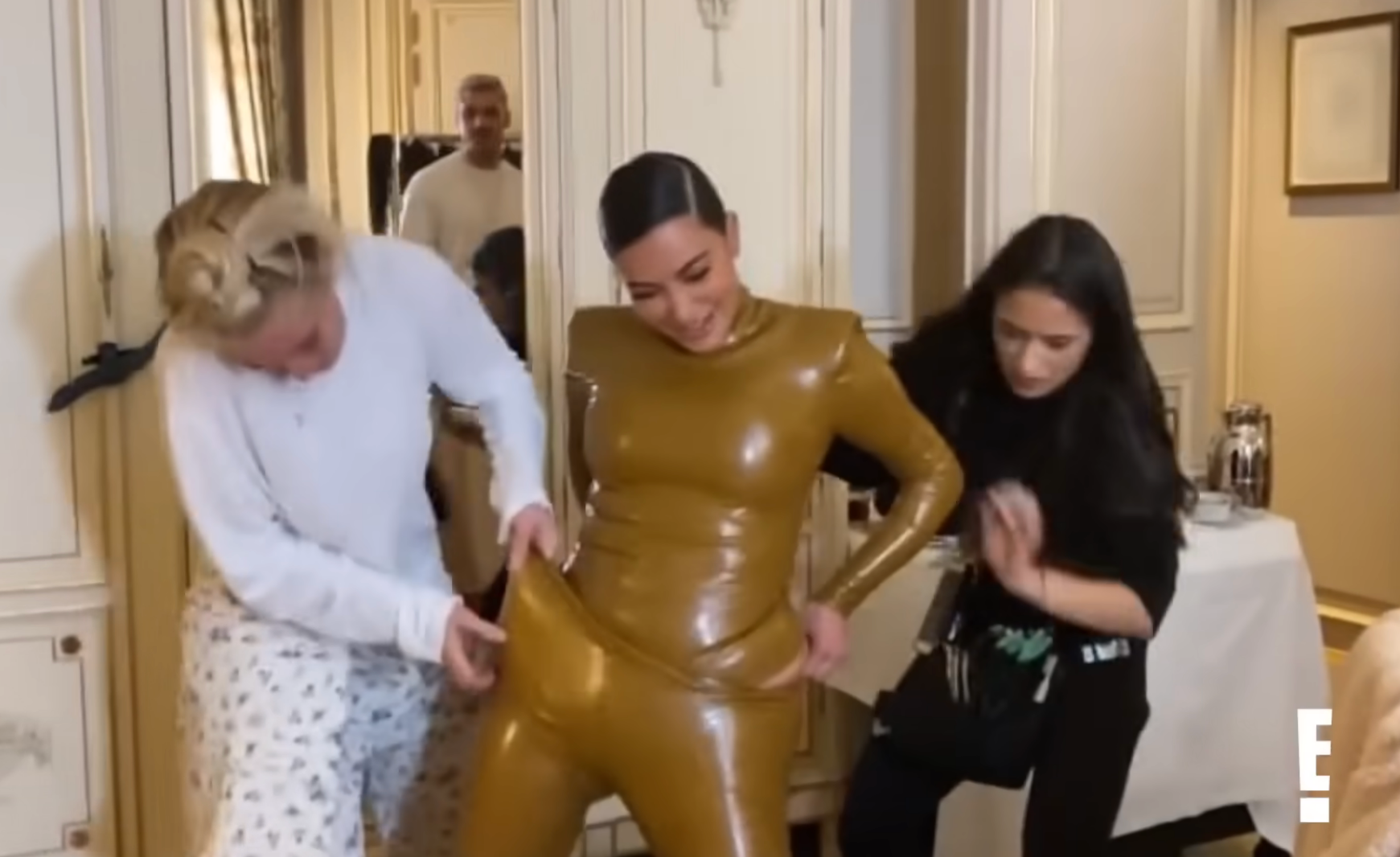 Two people help Kim adjust a shiny, form-fitting outfit