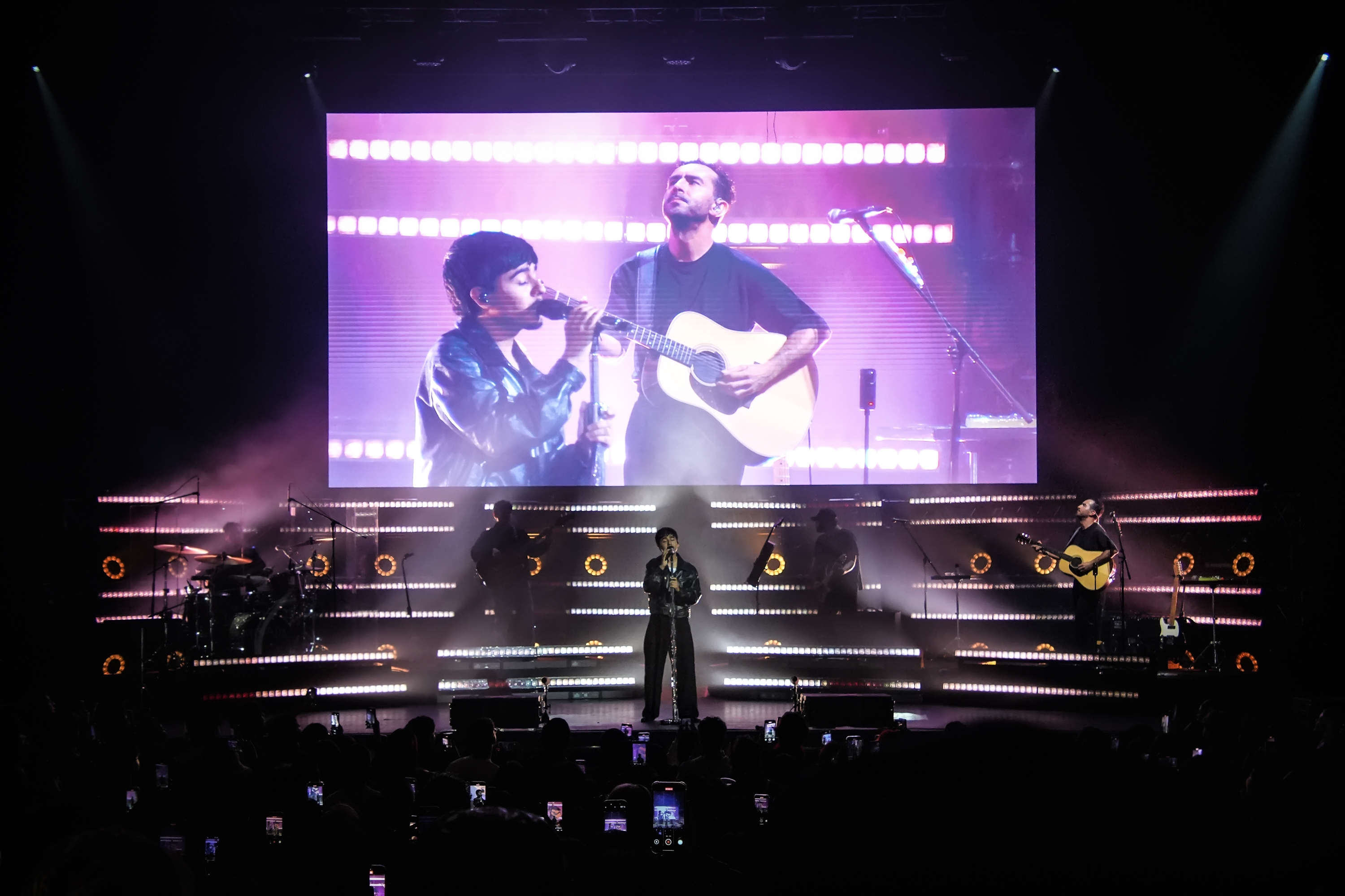 Concierto en vivo con banda en escenario. Un cantante en primer plano, otro músico tocando guitarra en pantalla grande detrás. Público asistiendo