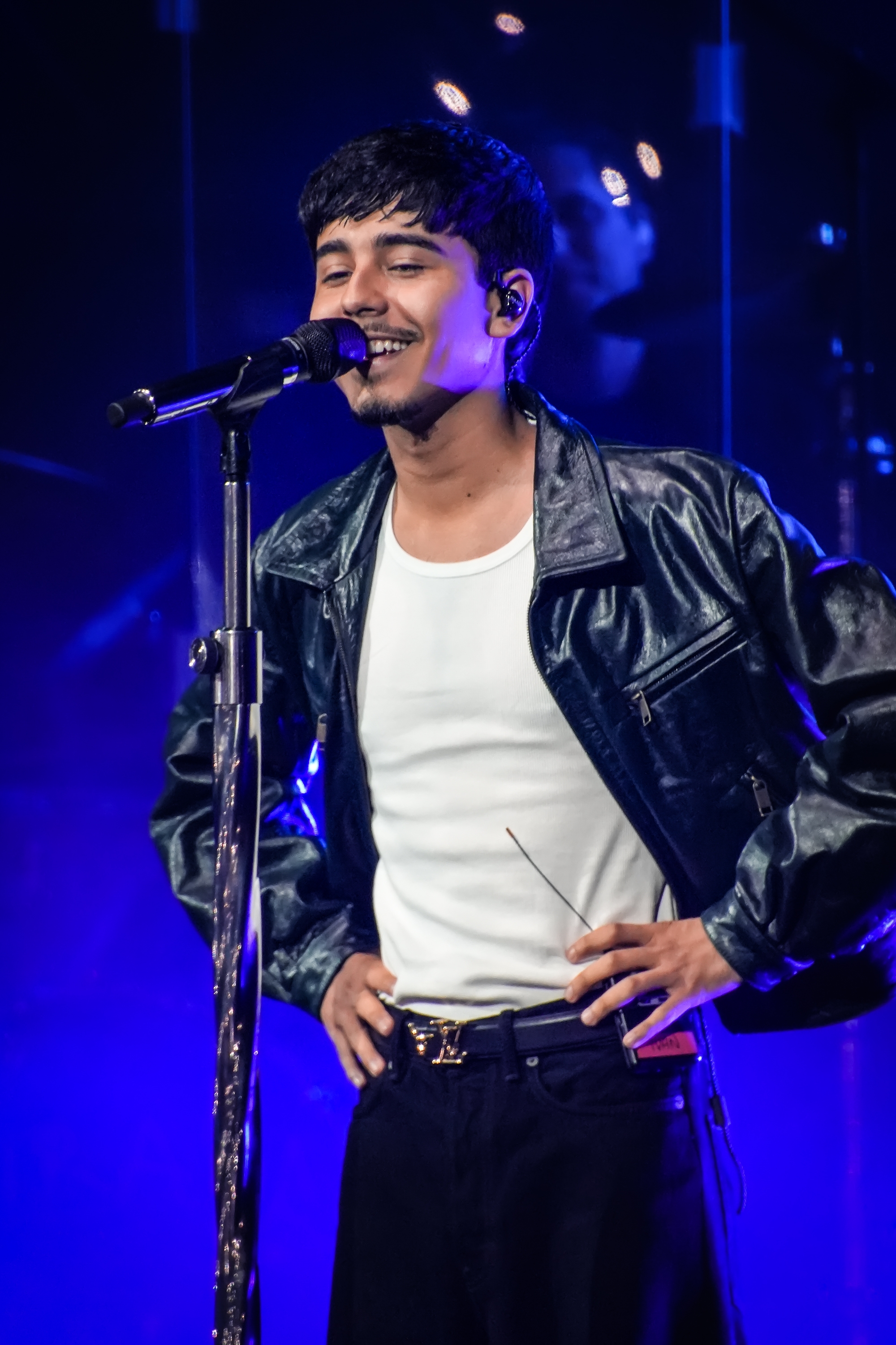 Persona cantando en el escenario, con chaqueta de cuero y camiseta blanca. Sostiene el micrófono y parece estar en un concierto