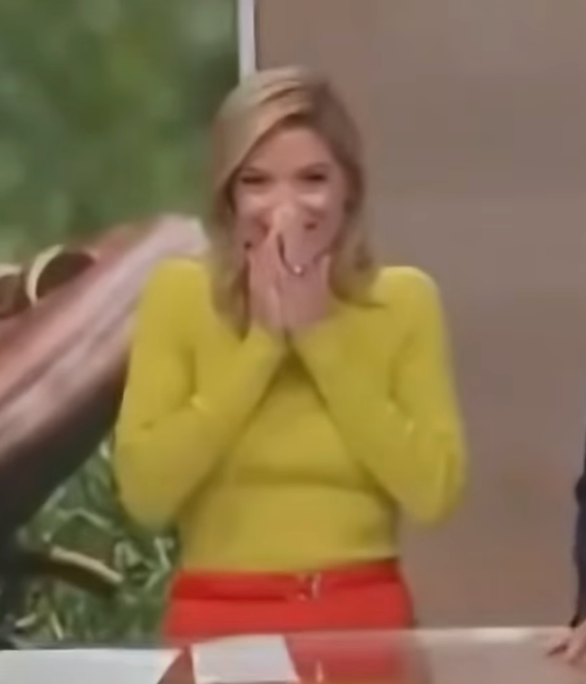A person standing and smiling with eyes closed, covering their mouth with their hands, wearing a long-sleeve top and skirt, appears delighted