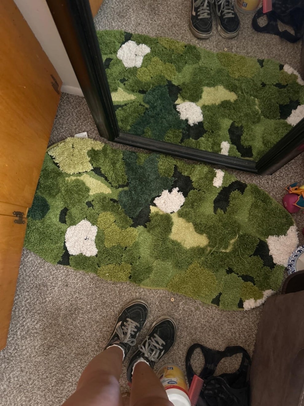 Person wearing sneakers stands on a green camouflage-patterned oval rug, reflected in a mirror