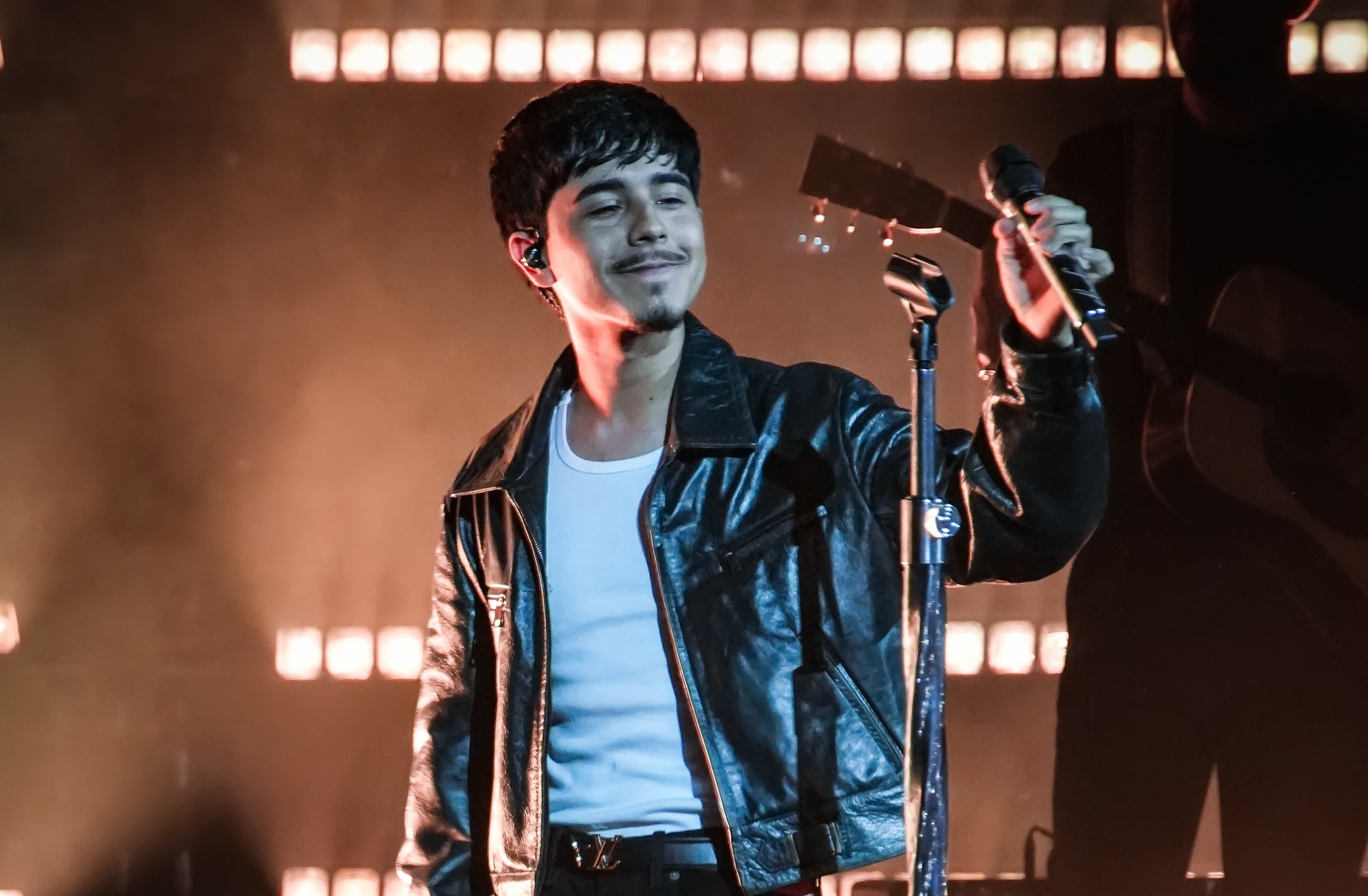 Cantante en concierto con chaqueta de cuero, camiseta y pantalones oscuros, sosteniendo un micrófono en un escenario iluminado