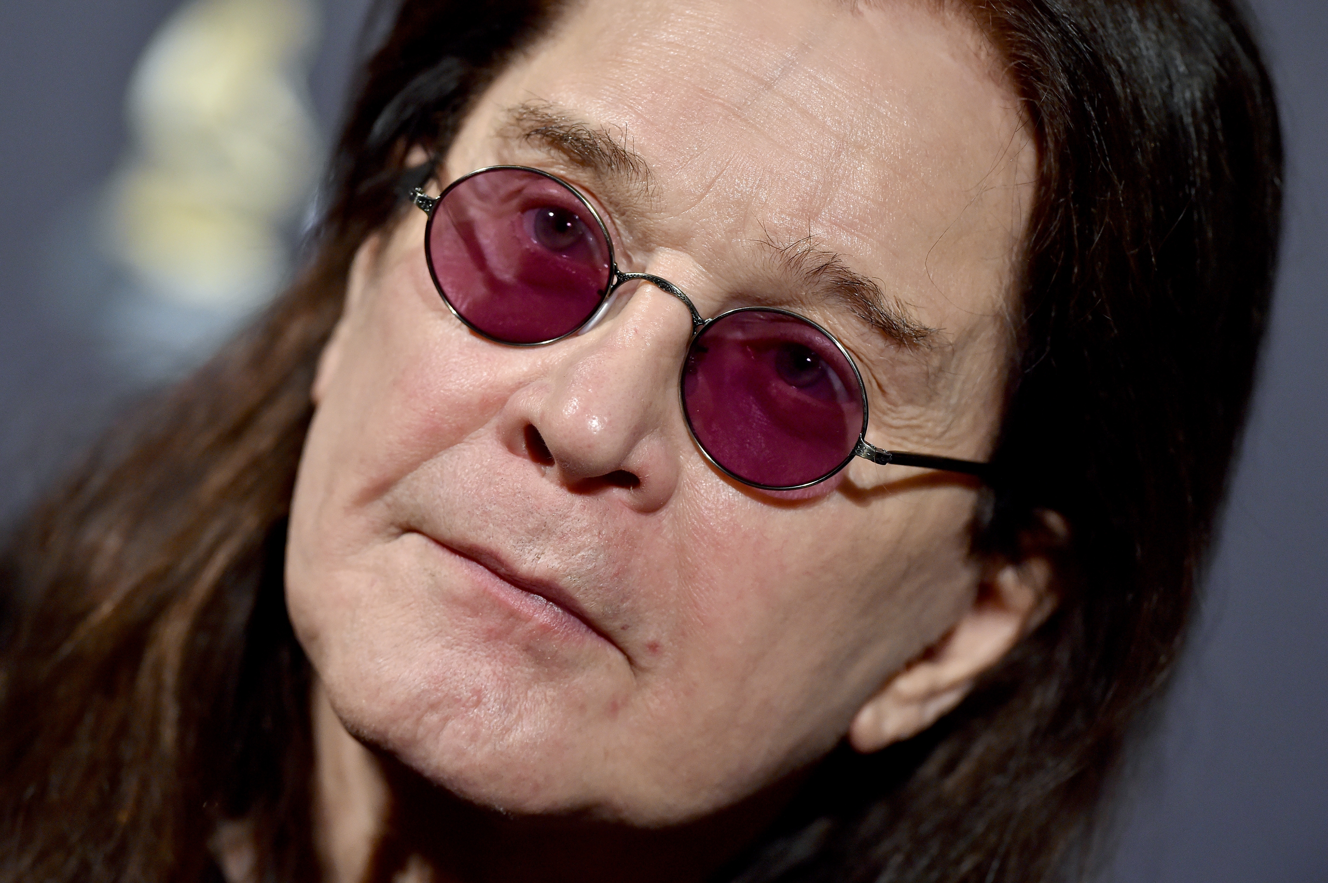 Ozzy Osbourne with long hair and round sunglasses looks towards the camera