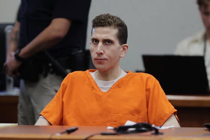 A person in an orange jumpsuit sits in a courtroom, looking forward. Two people in casual clothing are in the background