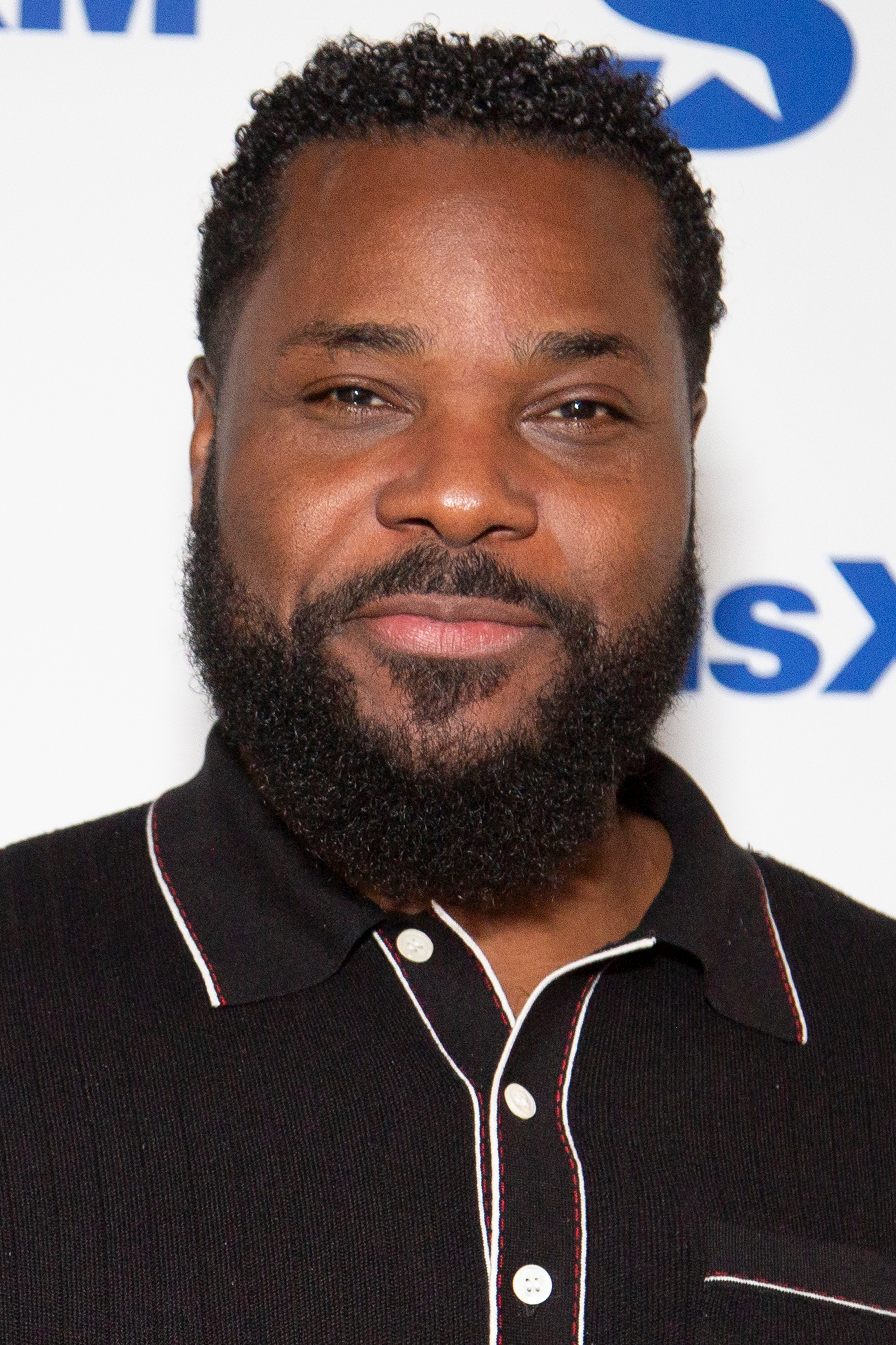 Malcolm Jamal Warner wearing a short-sleeved polo shirt with accents, smiling slightly at the camera