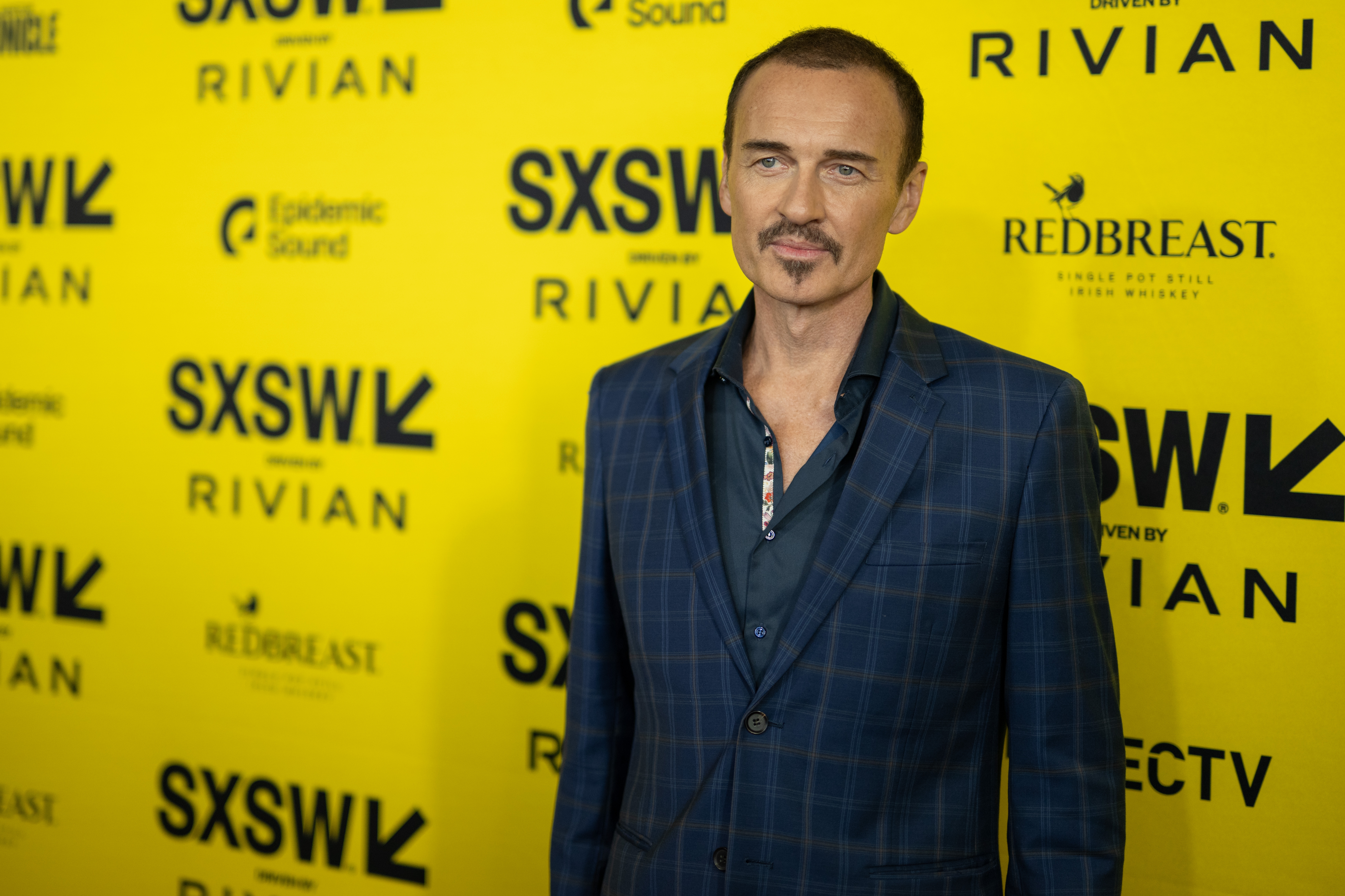 Julian McMahon in a plaid suit on a SXSW event backdrop, looking at the camera