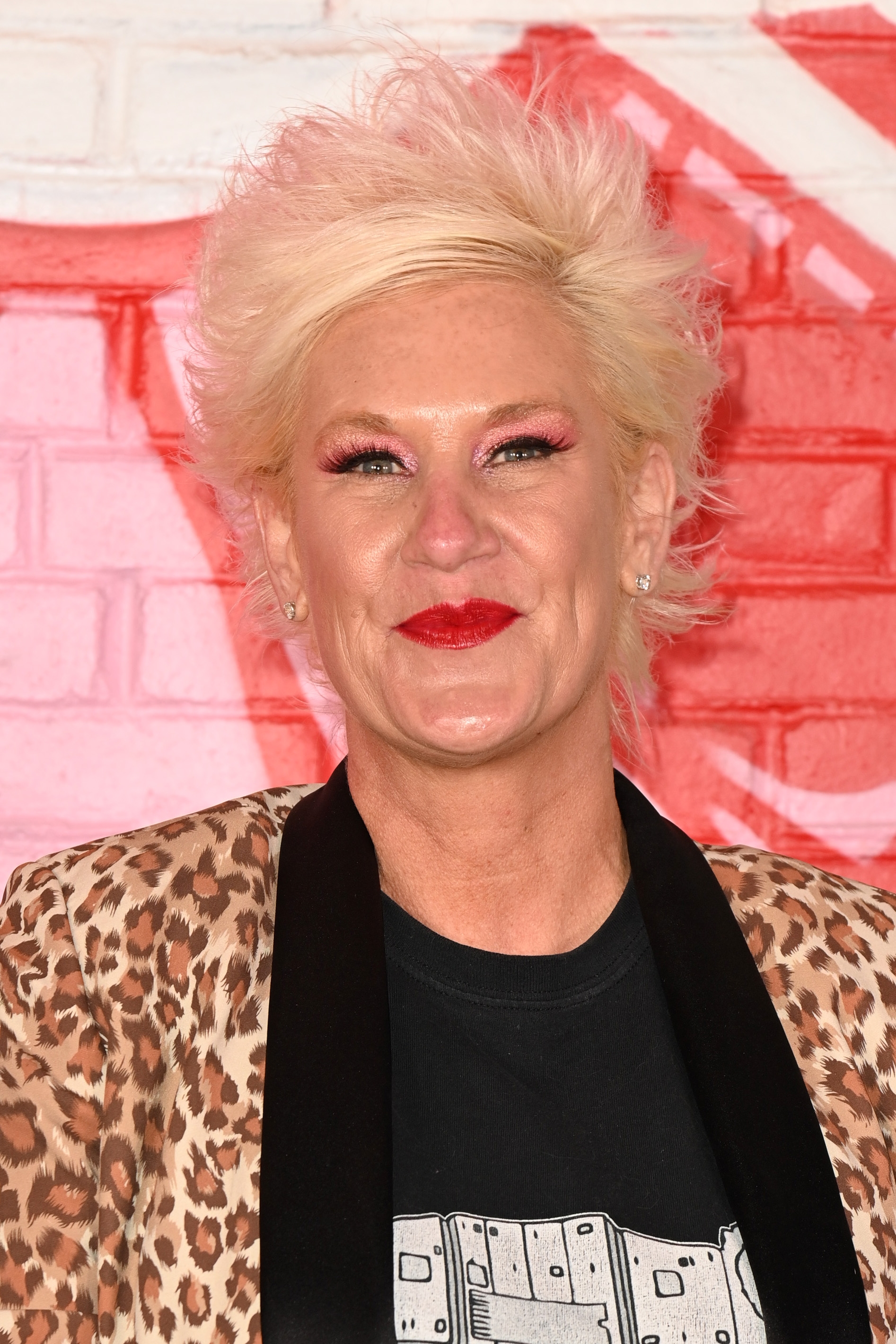 Anne Burrell with short, spiky hair, wearing a leopard print jacket over a graphic tee, smiles in front of a brick wall with graffiti