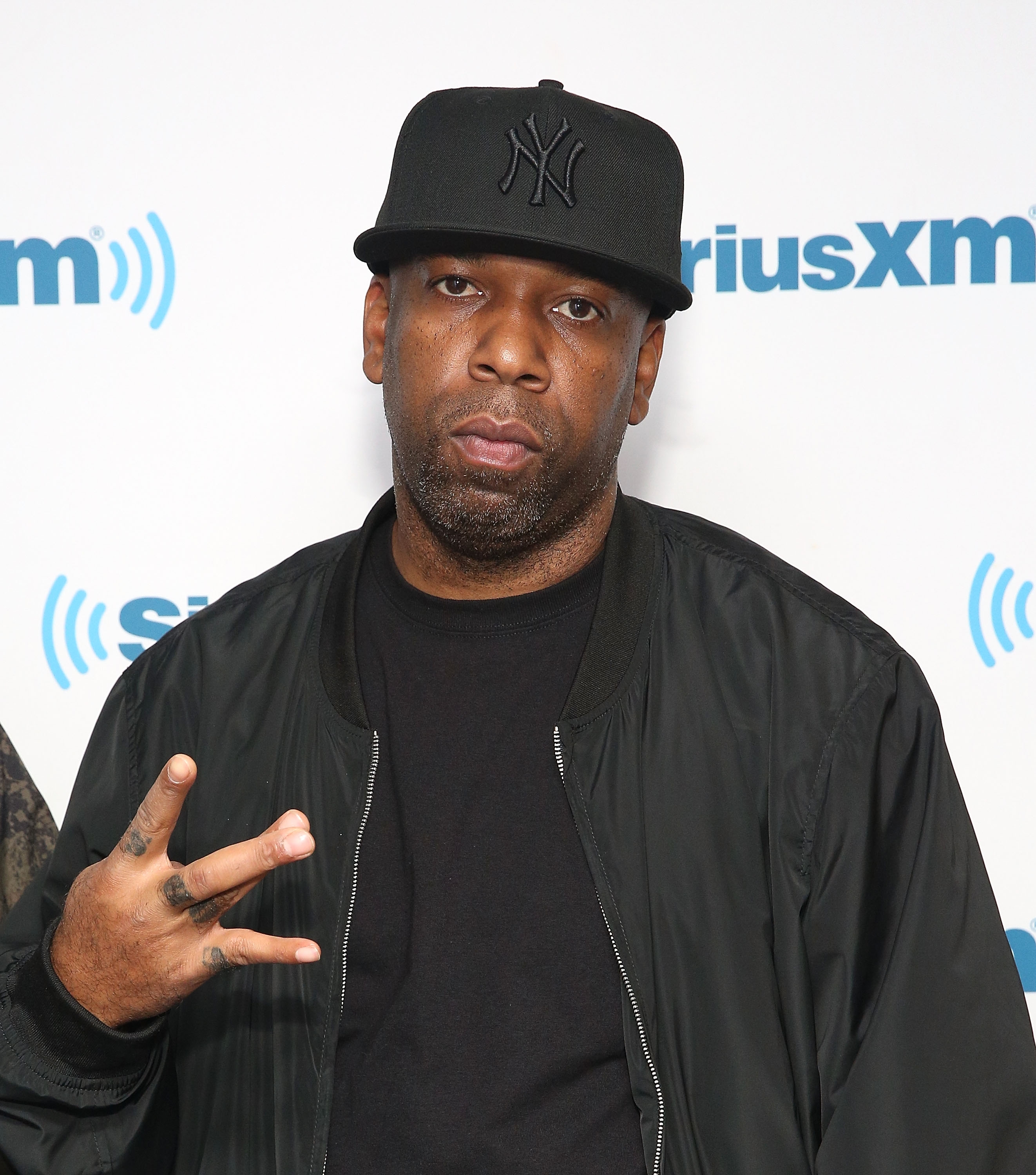 Young Noble from Outlawz in a black cap and jacket, posing with a hand gesture, in front of a SiriusXM backdrop