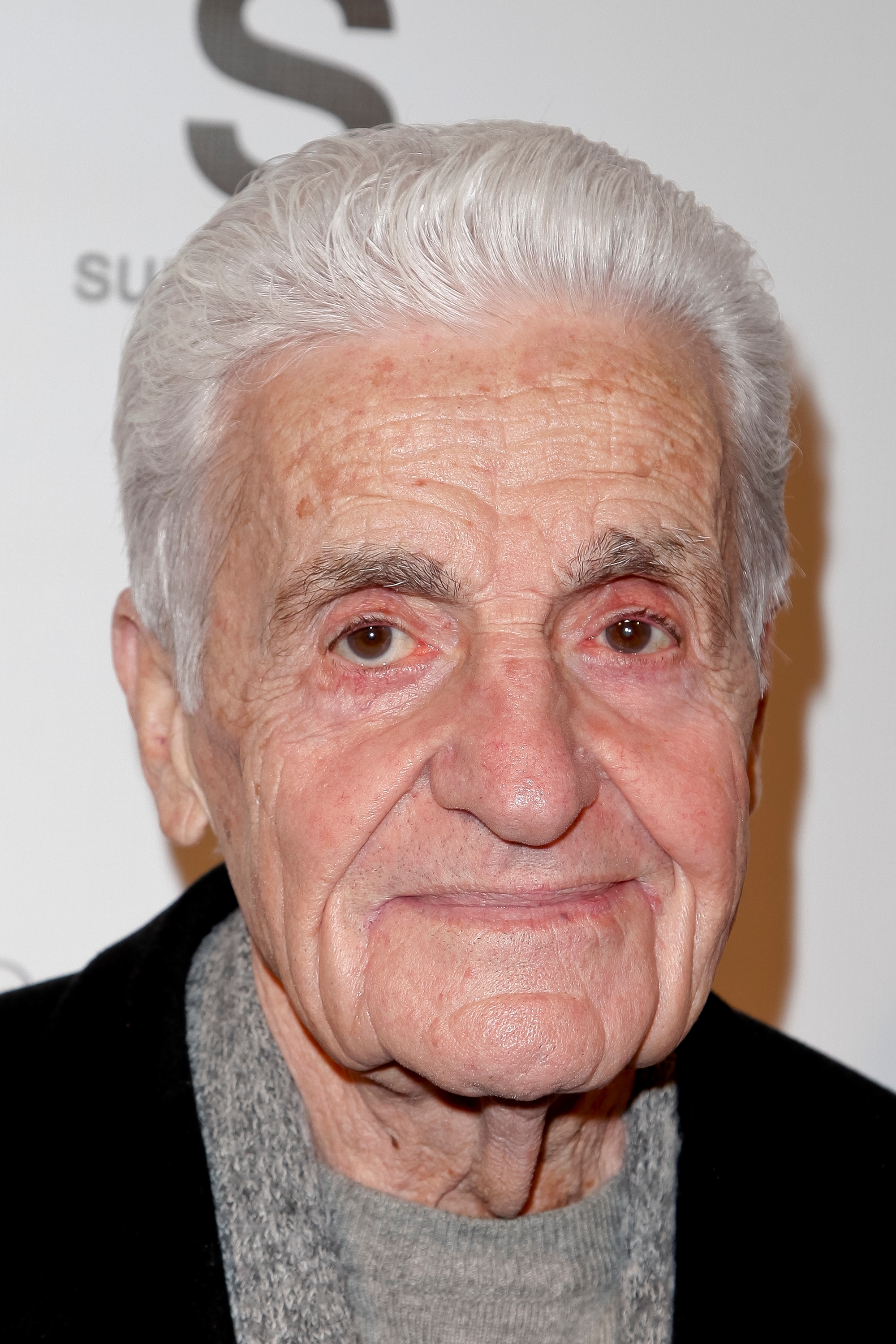 Older man with neatly styled white hair, wearing a dark jacket and gray sweater, posing in front of a step-and-repeat backdrop