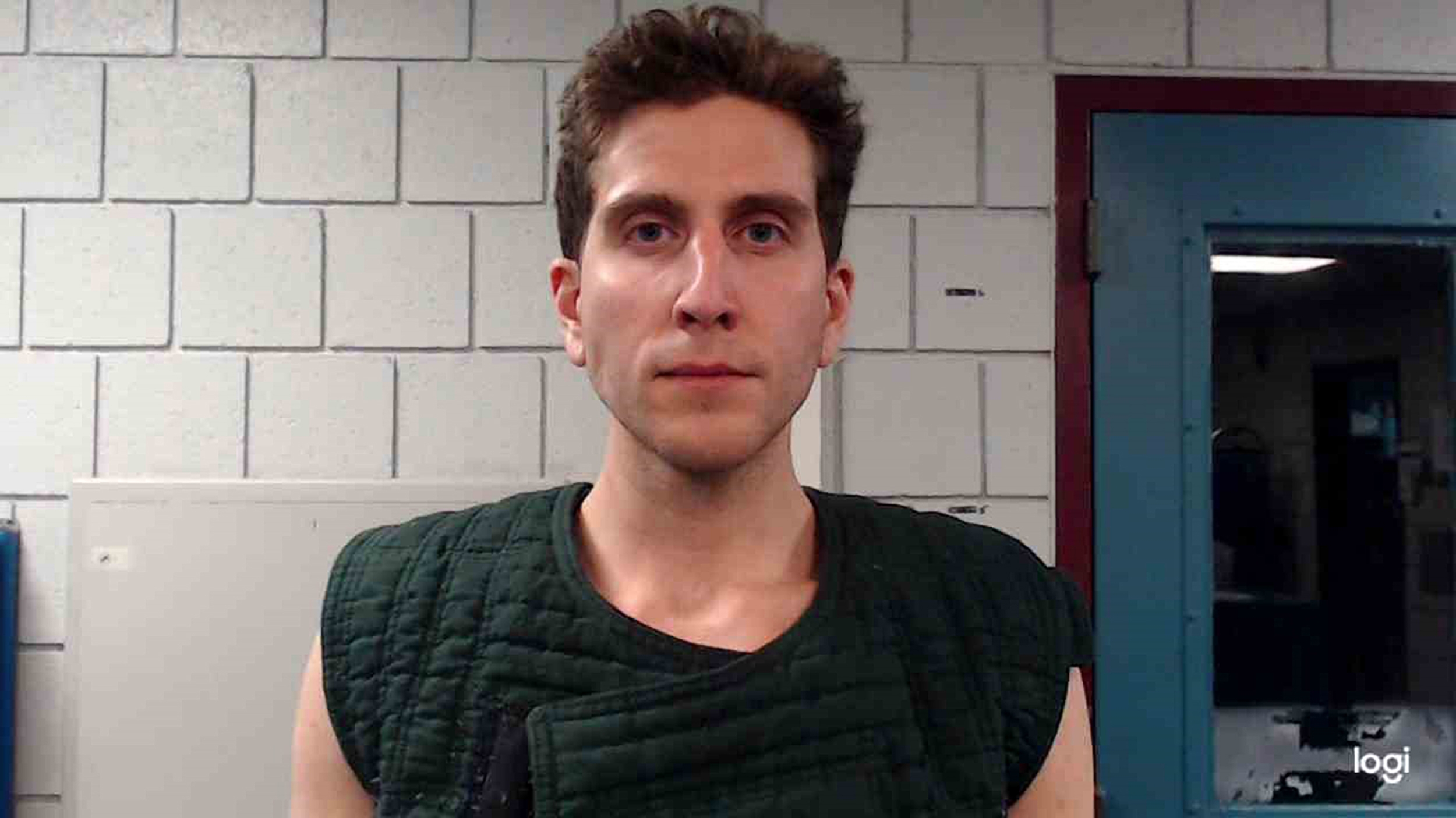 A person in a padded outfit stands in a room with a tiled wall, facing the camera with a neutral expression
