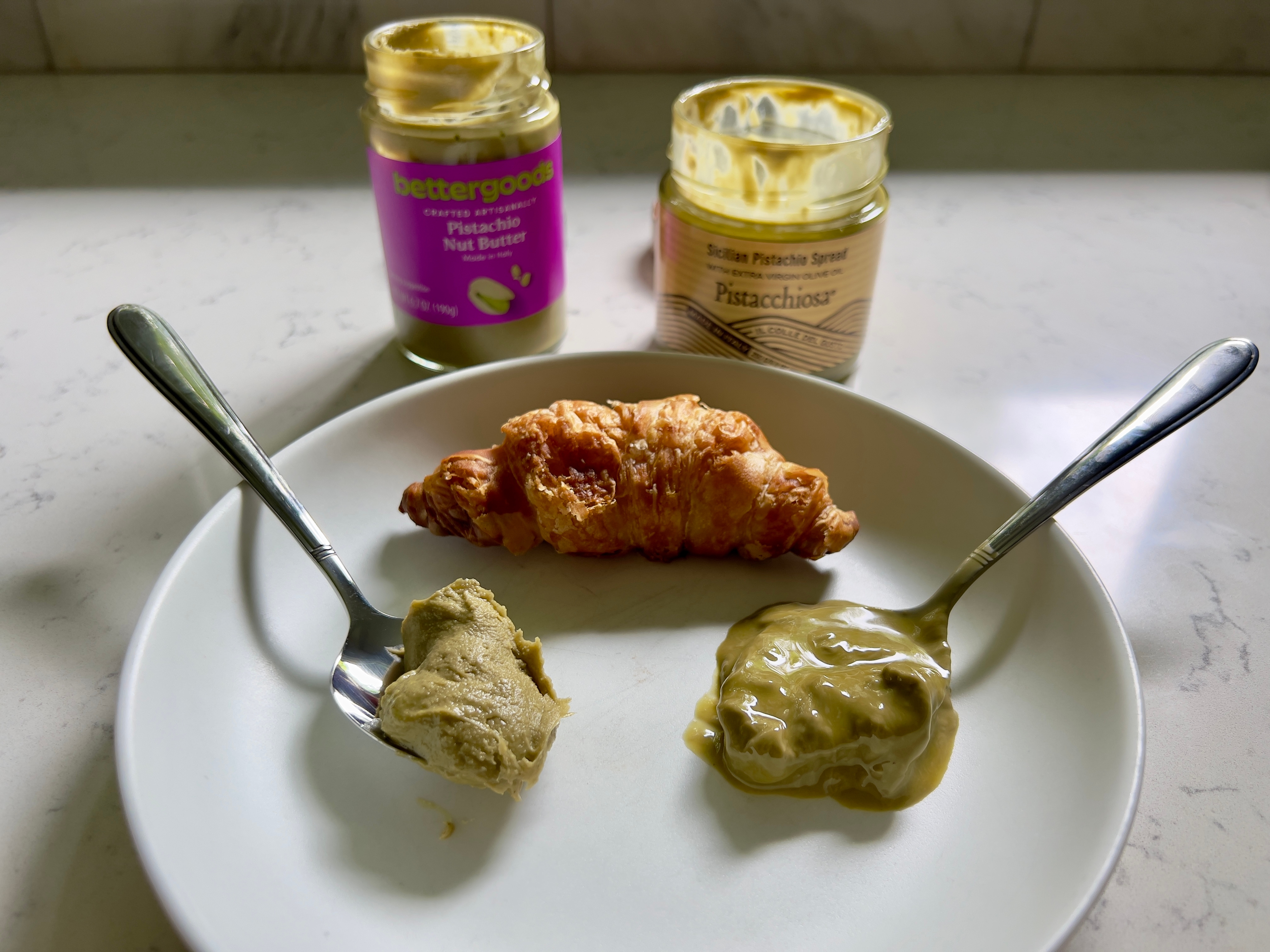 A croissant on a plate with two spoons of pistachio spreads beside it; jars of pistachio nut butter and cream behind