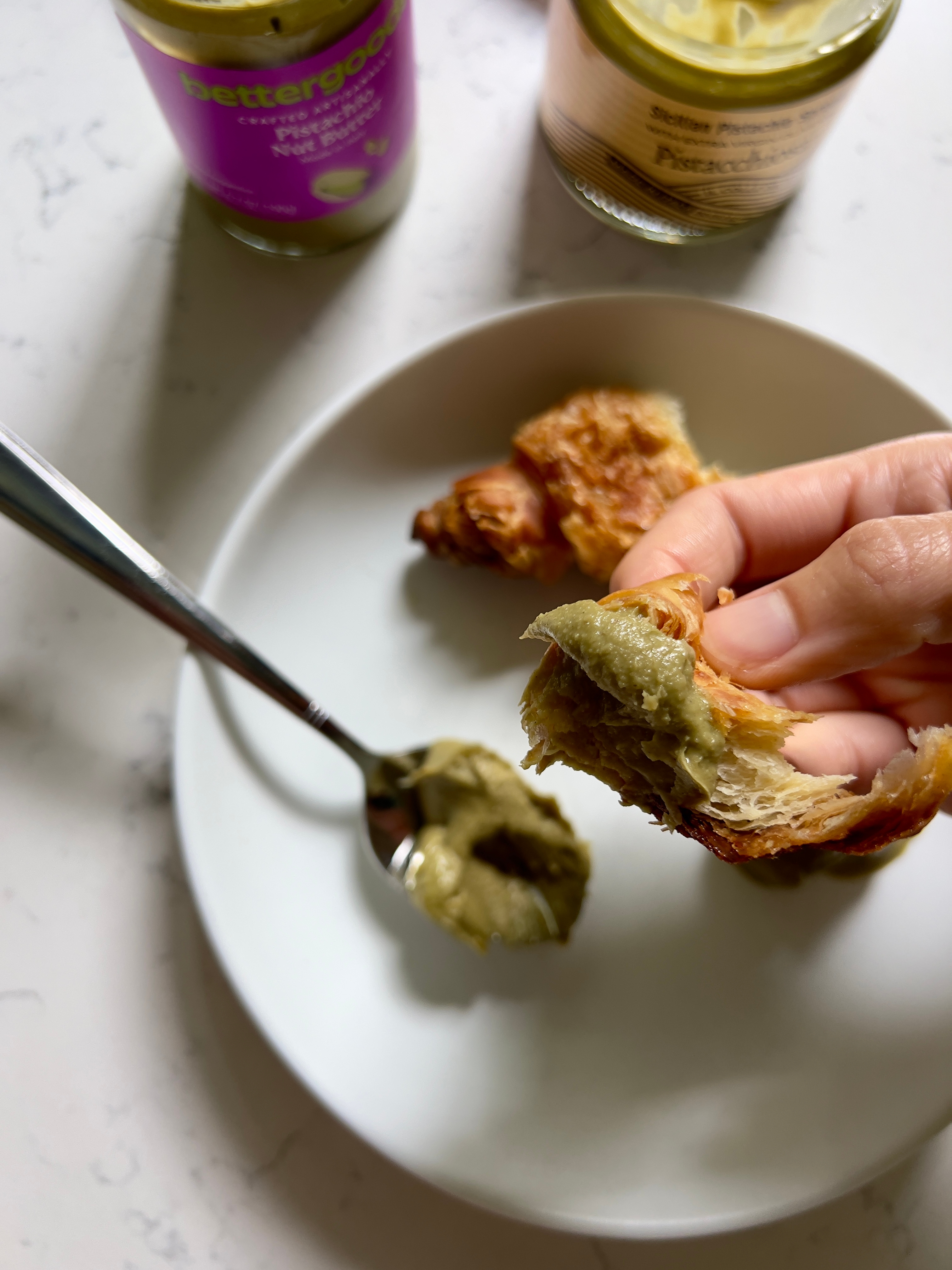 Hand holding a croissant piece with pistachio spread, spoonful on a plate; jars in the background