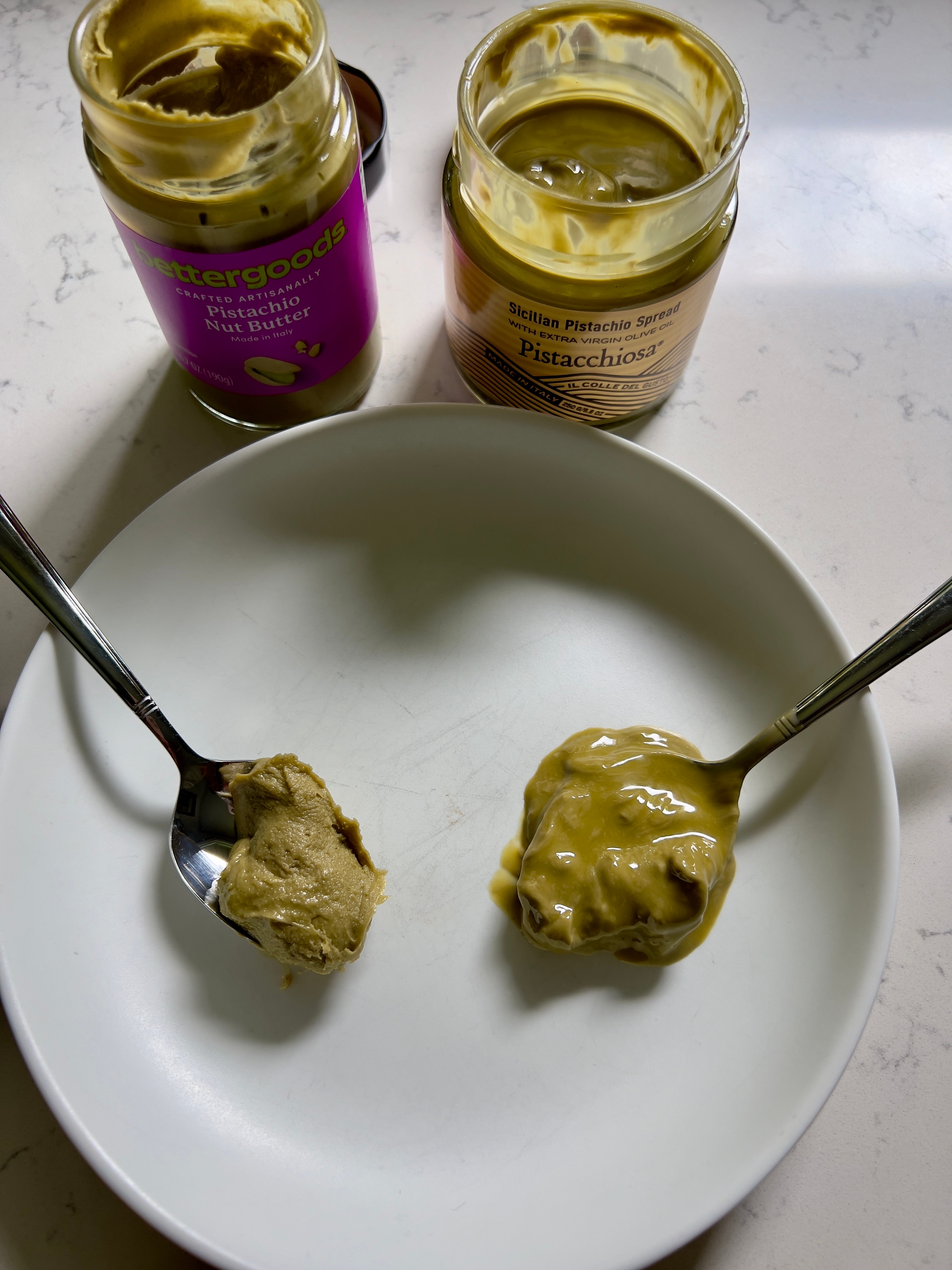Two spoons with different nut butters from jars are placed on a white plate, showcasing their textures for comparison