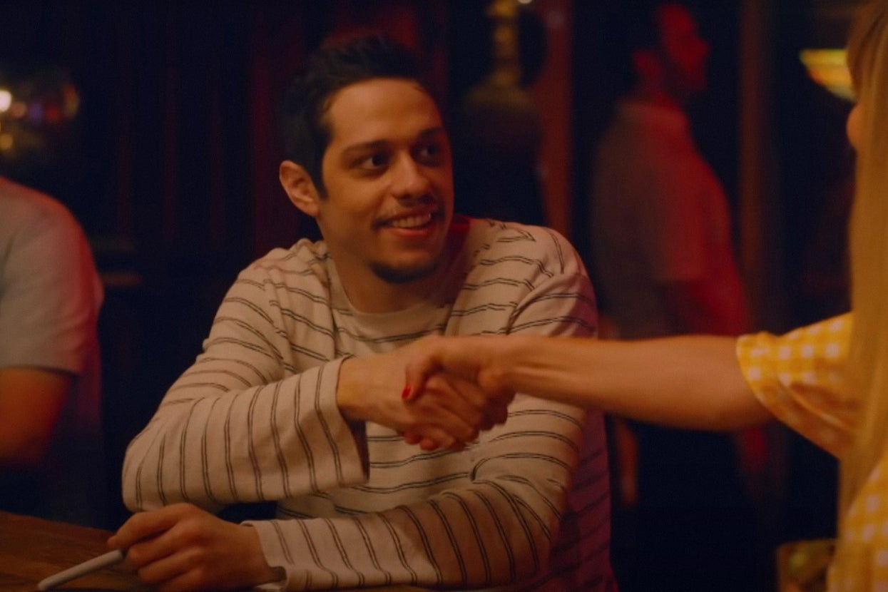 Pete Davidson in a striped shirt smiles while shaking hands with Kaley Cuoco in a yellow outfit at a dimly lit venue from "Meet Cute"