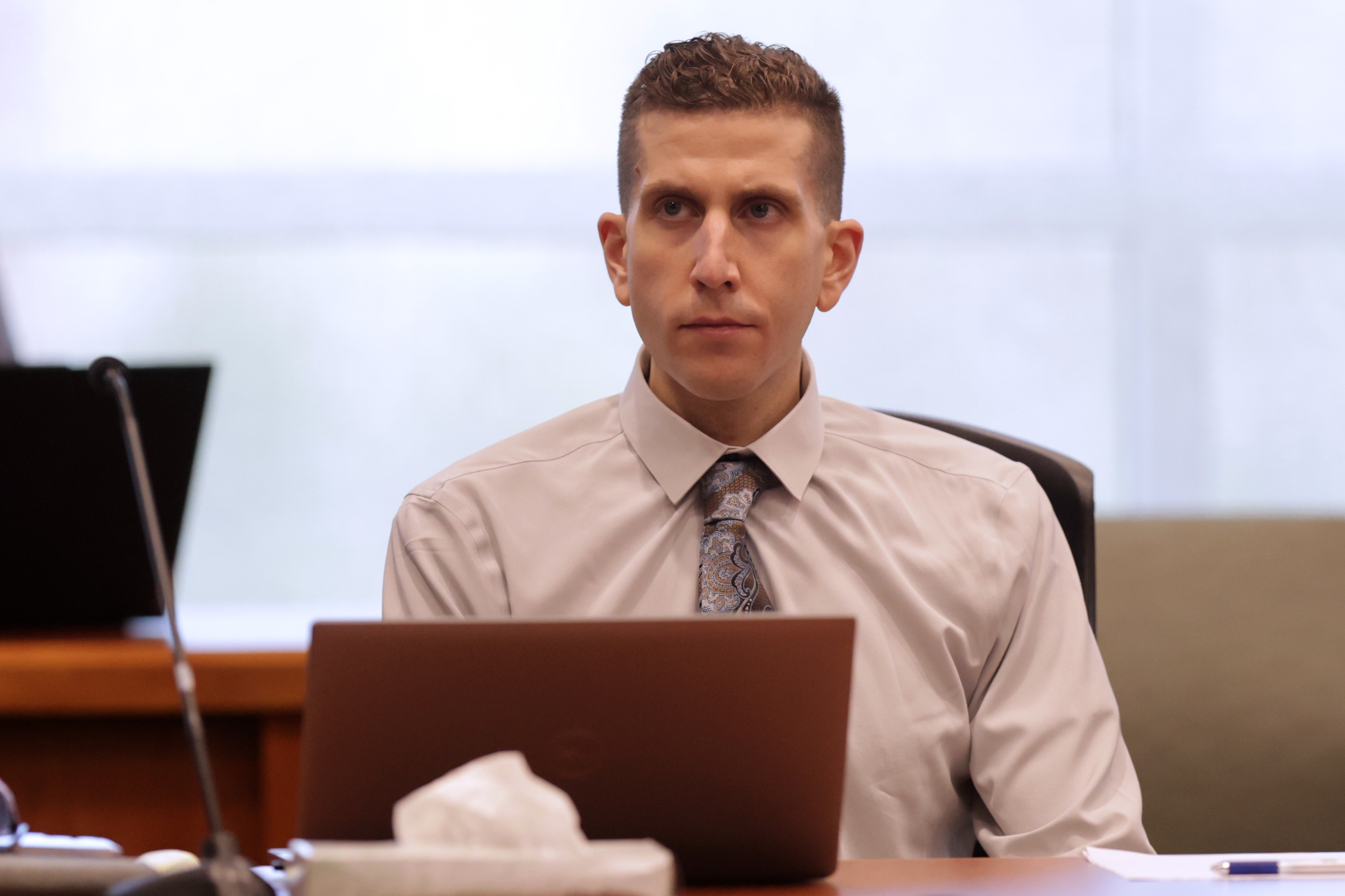Person in a formal shirt and tie sits at a table with a laptop in a courtroom setting, appearing focused