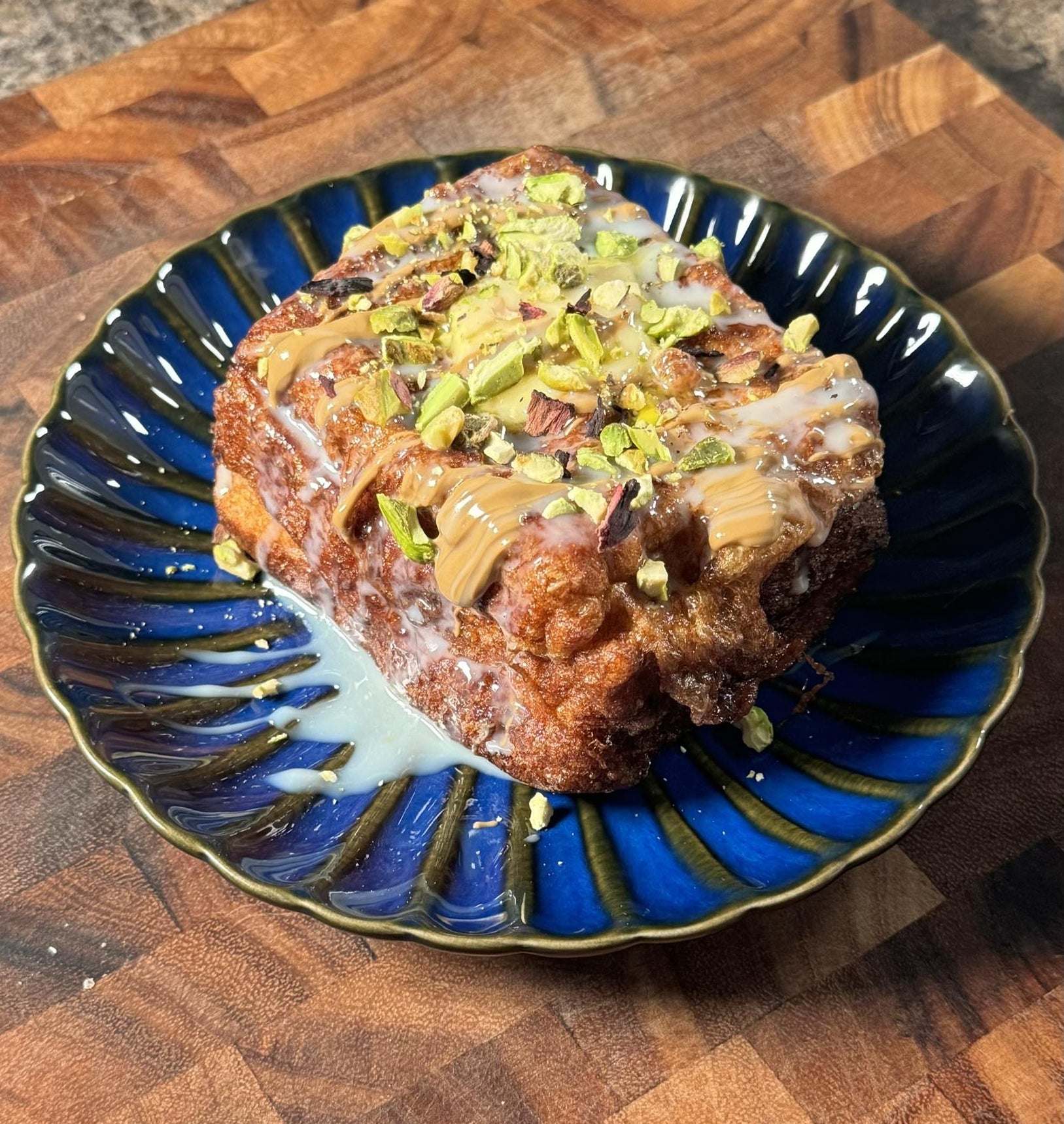 Pastry topped with drizzled icing and chopped pistachios on a decorative blue plate, placed on a wooden surface