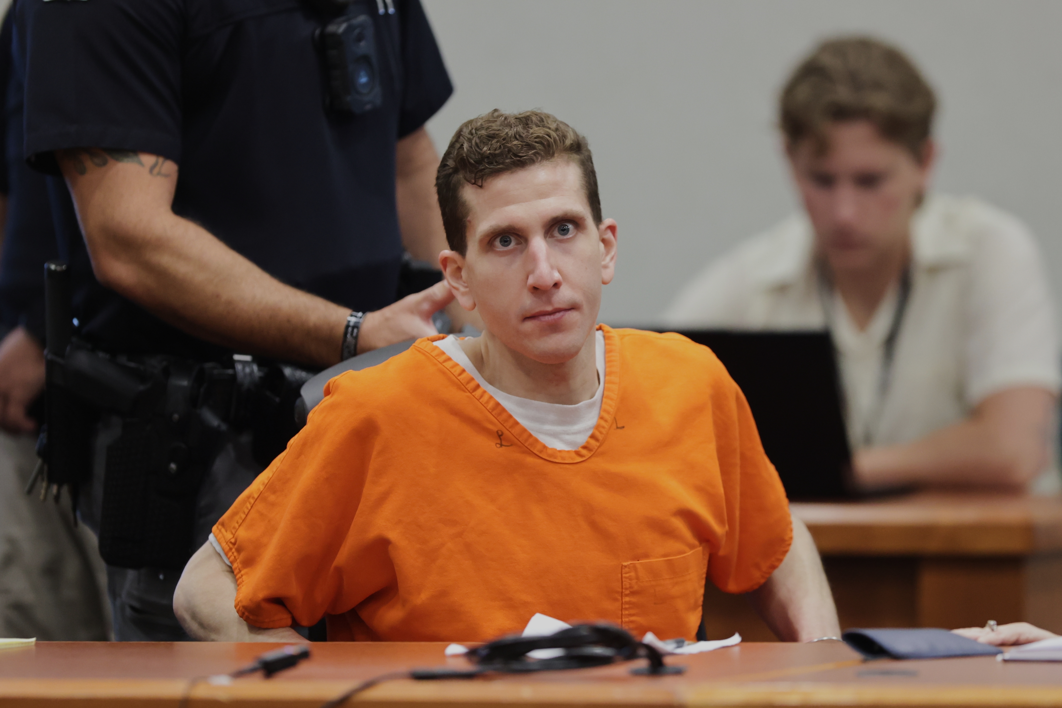 A man in an orange prison jumpsuit sits in a courtroom with a focused expression; a police officer stands nearby