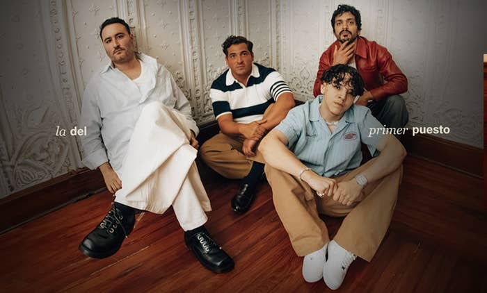 Cuatro hombres sentados en el suelo de una habitación elegante, uno con camisa blanca y pantalones claros, otro a rayas, otro rojo y el último azul