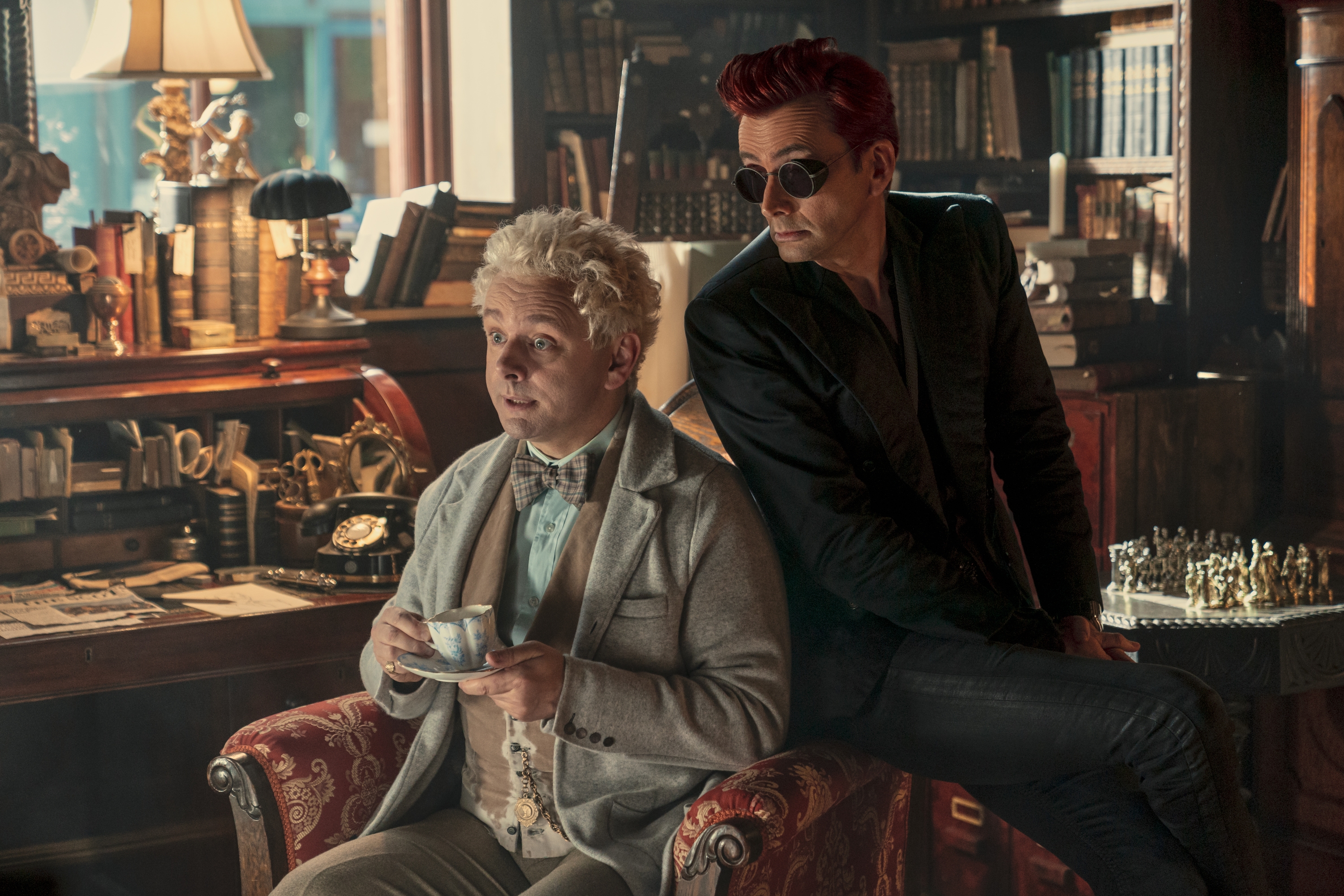 Dos actores en una escena de biblioteca: uno sentado con taza de té y el otro apoyado en el sillón, usando gafas de sol y traje oscuro