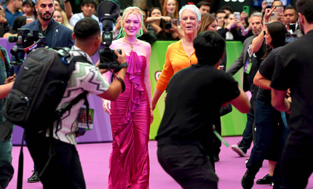 Dos personas sonrientes en un evento, una en vestido elegante, la otra en atuendo casual, rodeadas de fotógrafos y asistentes en alfombra