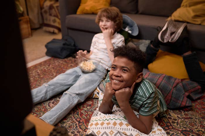 Two children smiling and watching TV while lying on the floor with popcorn. Another child is stretched upside down on the couch behind them