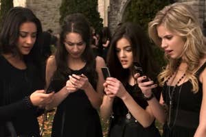 Four people in stylish black outfits intently look at their phones, standing outdoors with a stone wall backdrop